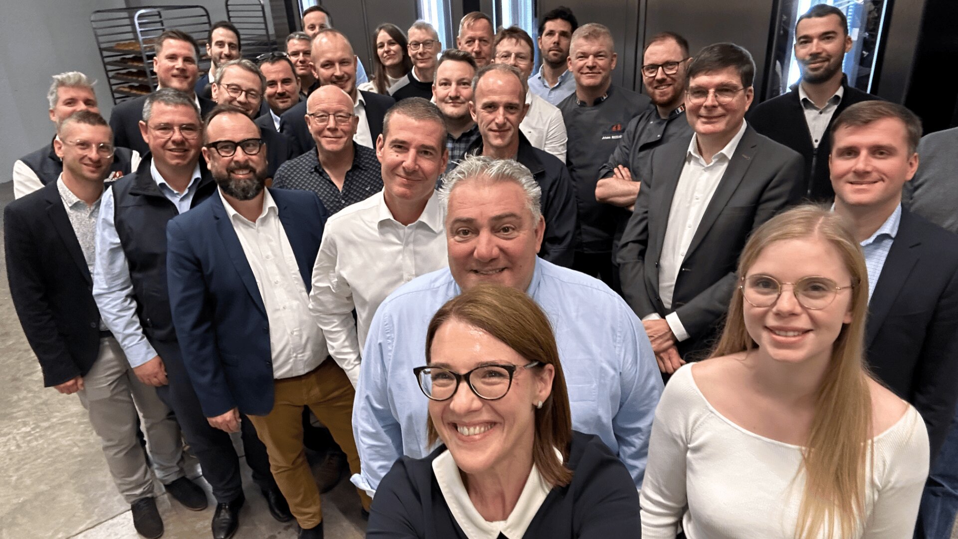 A group of about 25 people, dressed in business casual attire, smile and pose together for a group selfie indoors, with tables and metal shelving visible in the background.