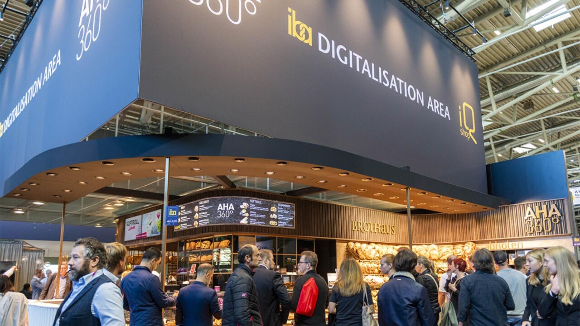 A busy exhibition booth labeled Digitalisation Area and AHA 360°, with people gathered around, some interacting and others looking at bakery products displayed on shelves. The setting appears modern and professional.