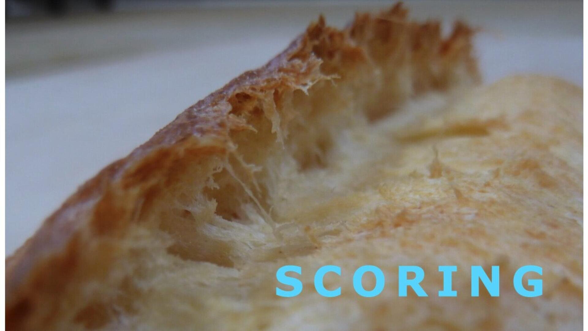 Nahaufnahme des knusprigen Randes eines gebackenen Brotes, der seine luftige, faserige Textur zeigt. Das Wort SCORING erscheint in großen hellblauen Buchstaben am unteren rechten Bildrand.
