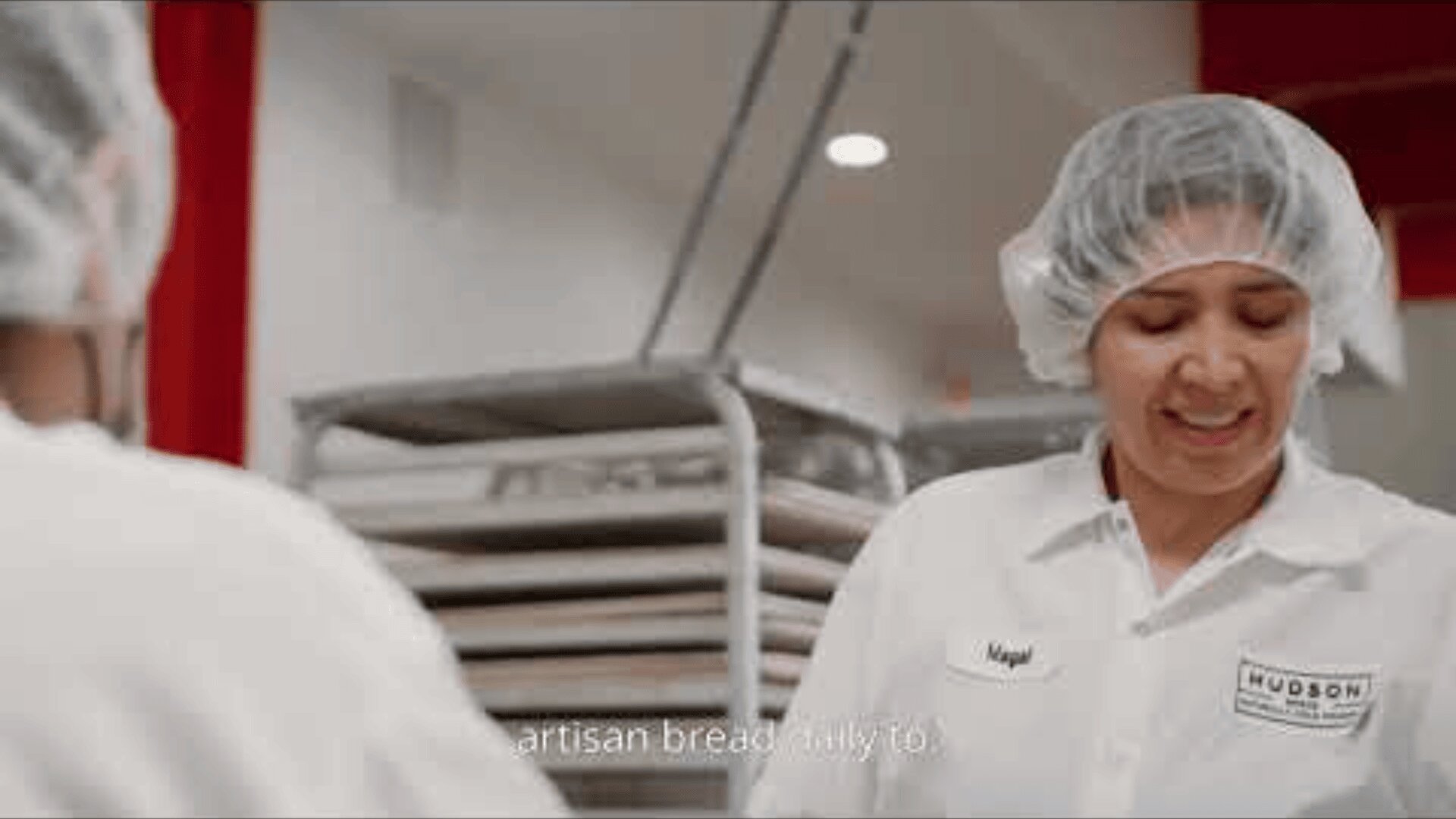 Eine Bäckereimitarbeiterin in weißer Uniform und Haarnetz lächelt bei der Arbeit, während sich im Hintergrund Tabletts stapeln. Auf dem Namensschild der Arbeiterin steht "Magal".