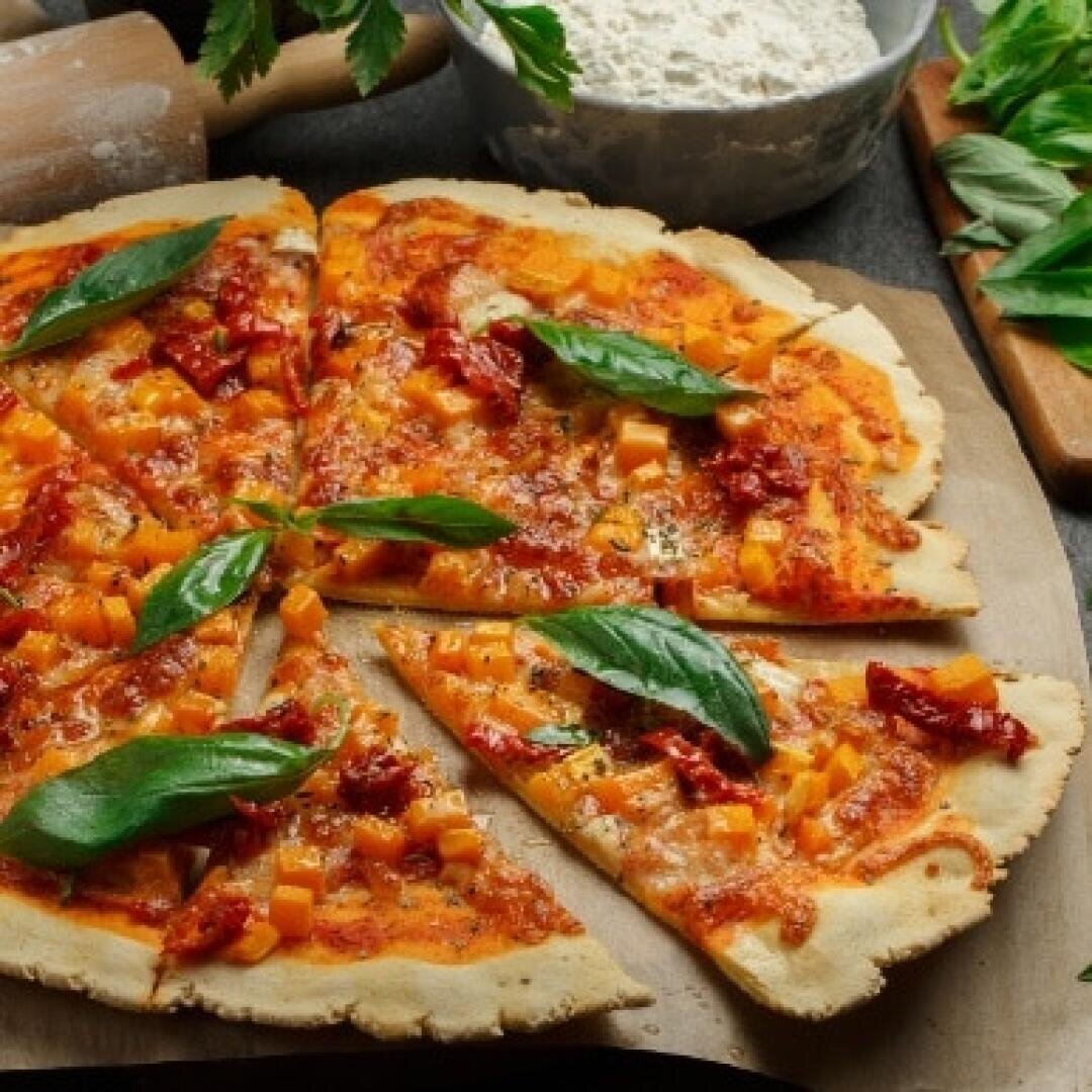 A freshly baked thin-crust pizza topped with melted cheese, diced tomatoes, sun-dried tomatoes, and fresh basil leaves sits on parchment paper, surrounded by baking ingredients and tools.