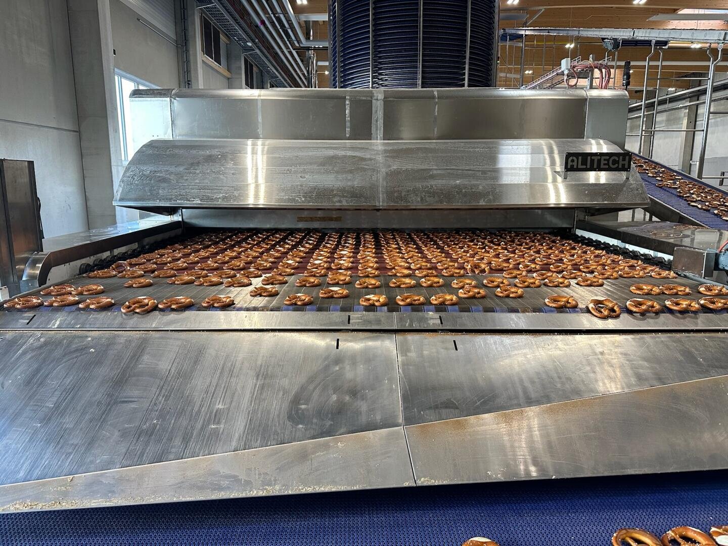 A large industrial oven conveyor bakes many pretzels in a modern food processing facility, with pretzels lined up neatly on metal trays.
