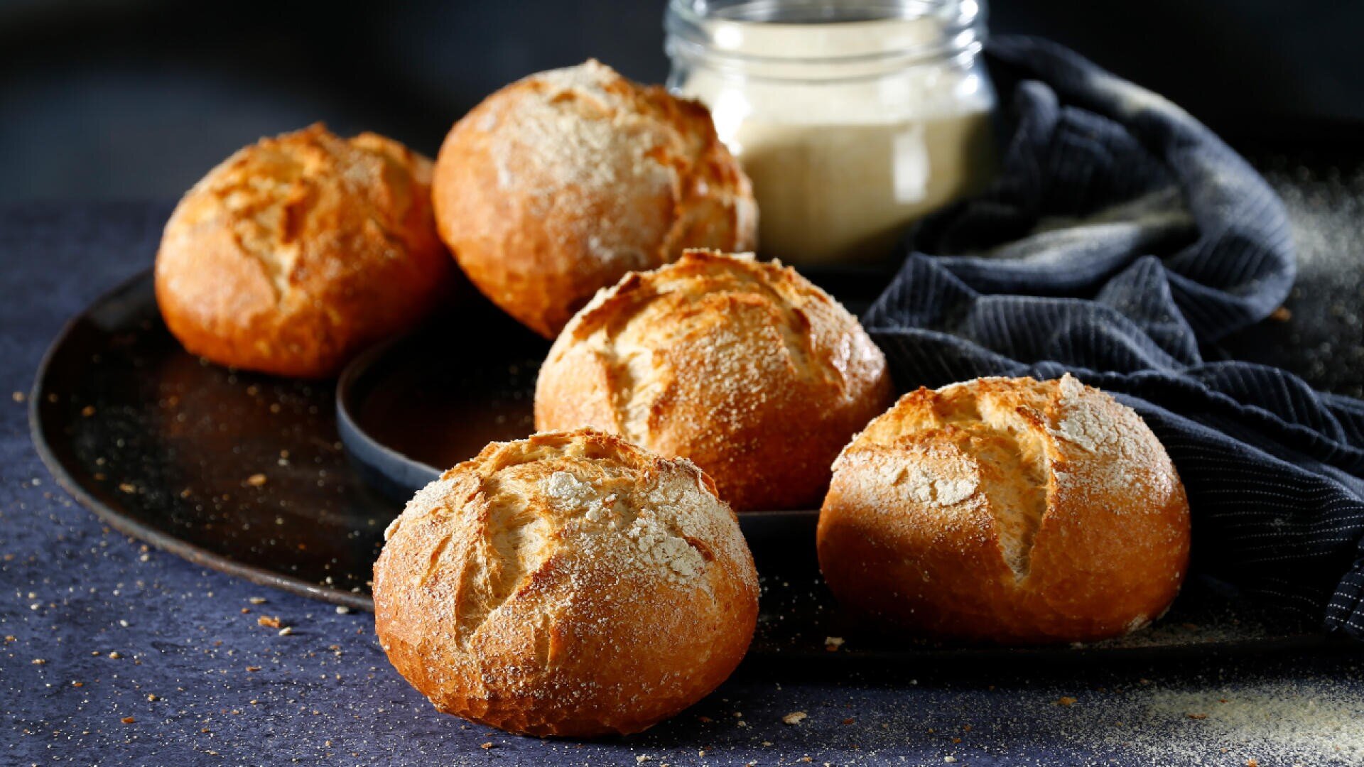 Fünf runde, knusprige Brötchen liegen auf einem dunklen, mit Mehl bestäubten Tablett. Im Hintergrund stehen ein Glasgefäß und ein dunkel gestreiftes Tuch, die für eine rustikale Küchenatmosphäre sorgen.