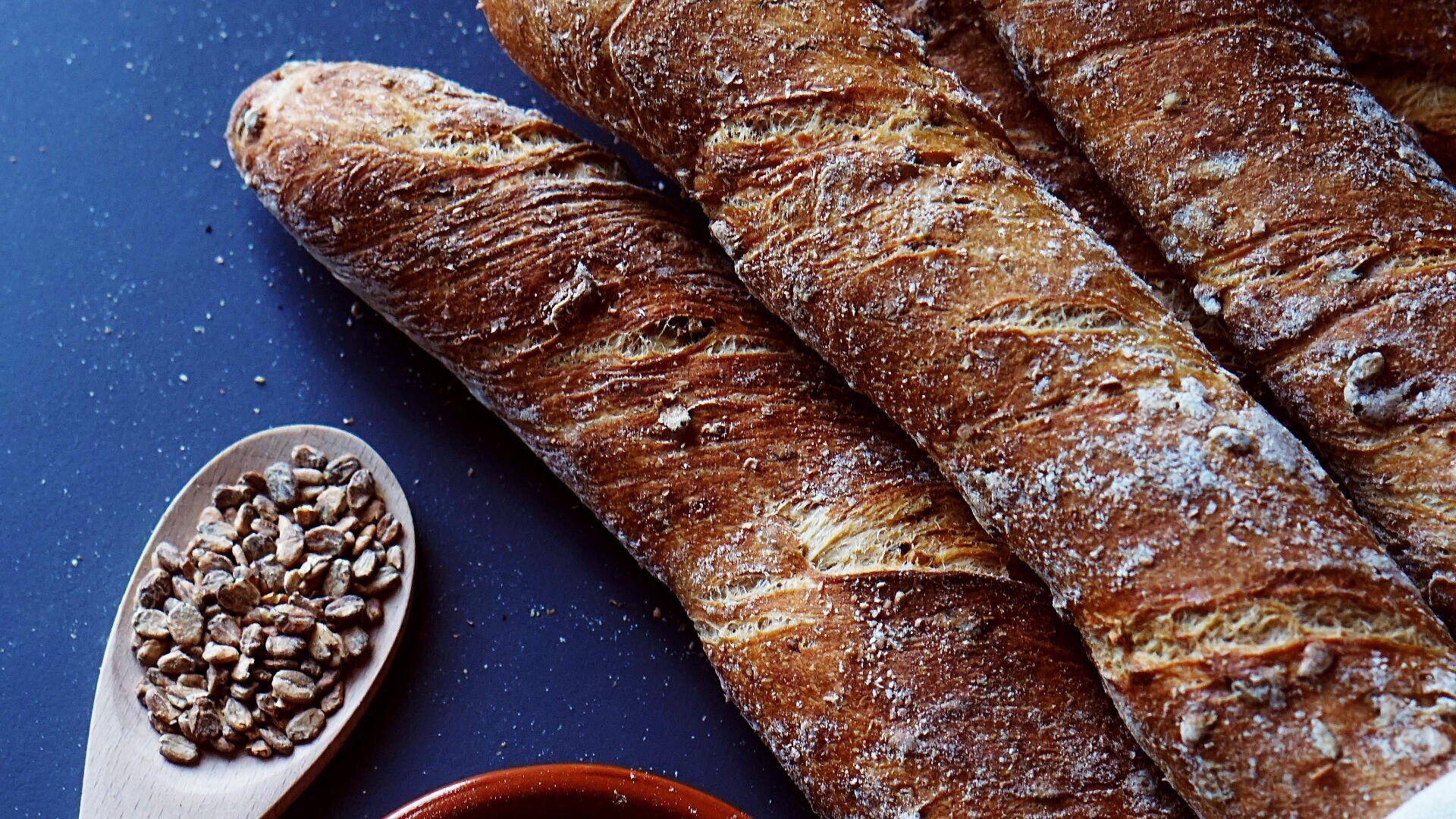 Three crispy, golden-brown baguettes rest on a dark surface beside a wooden spoon filled with seeds and a partial view of a small red dish.