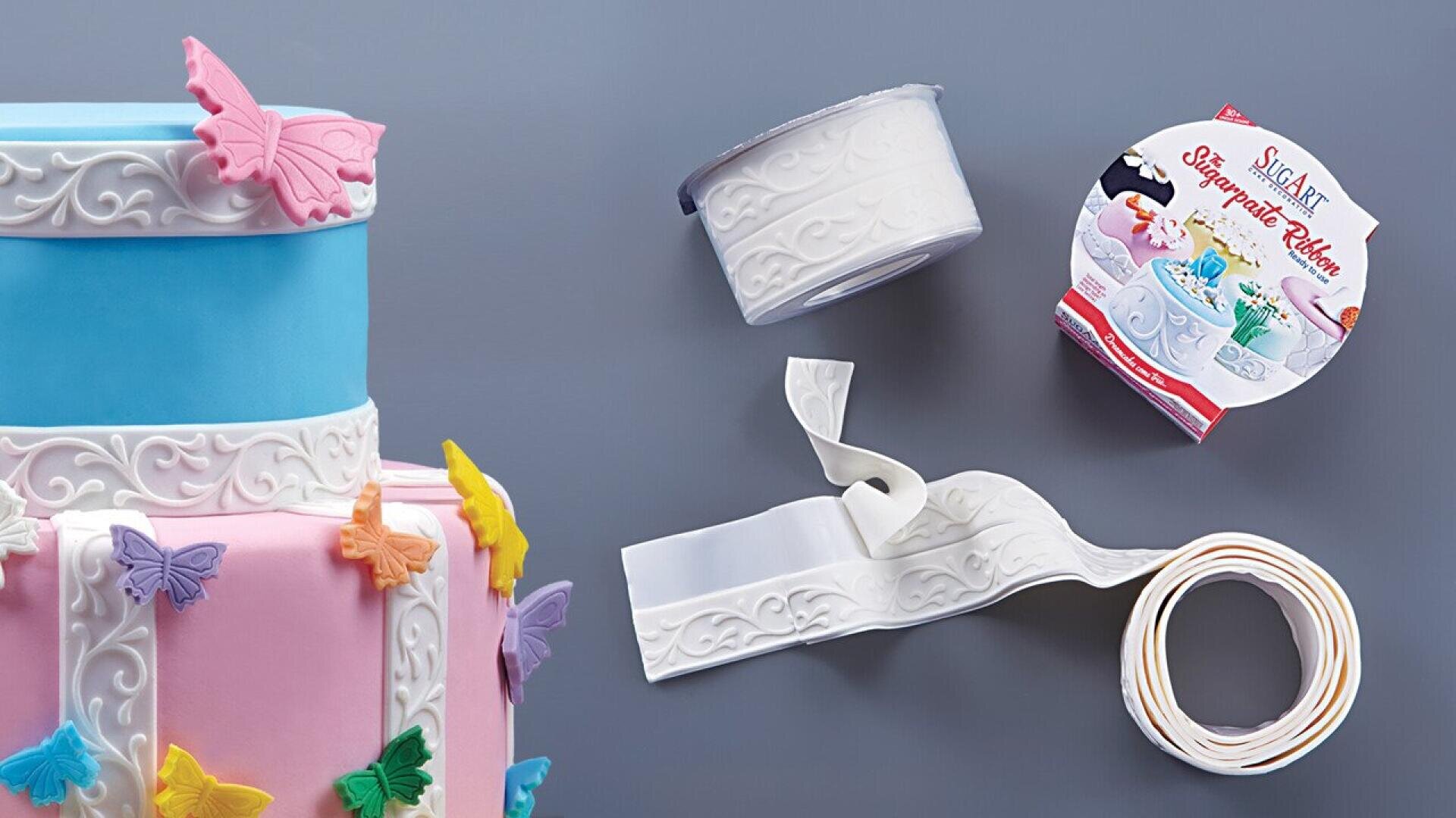 A partial view of blue and pink cakes decorated with colorful butterflies and embossed white sugar ribbon. Nearby, a roll of the same sugar ribbon and its packaging are shown on a gray surface.