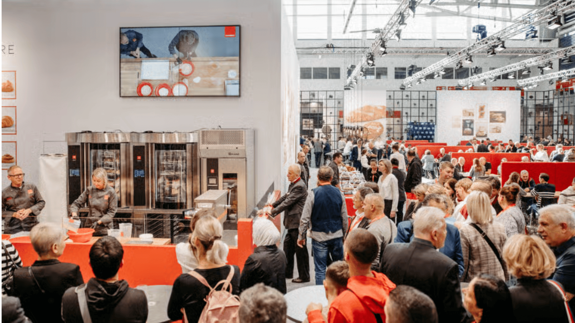 Eine große Menschenmenge verfolgt eine Kochvorführung auf einer Fachmesse. Zwei Personen stehen hinter einer Theke mit Backgeräten, während andere zusehen. Auf einem großen Bildschirm wird eine Nahaufnahme der Vorführung gezeigt.