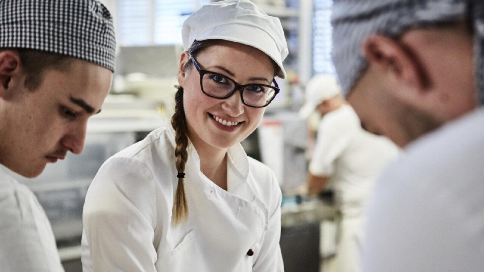 A woman in a white chef’s uniform and cap smiles at the camera while working with two other chefs in a busy kitchen. The chefs are focused on preparing food.