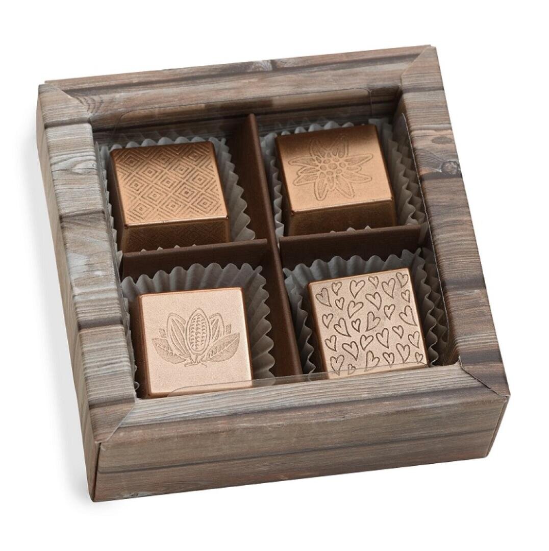 Eine Holzschachtel mit vier quadratischen Pralinen, jede in einem separaten Fach. Auf den Pralinen sind verschiedene Motive eingraviert: geometrische Formen, eine Blume, eine Kakaoschote und ein Muster aus Herzen.