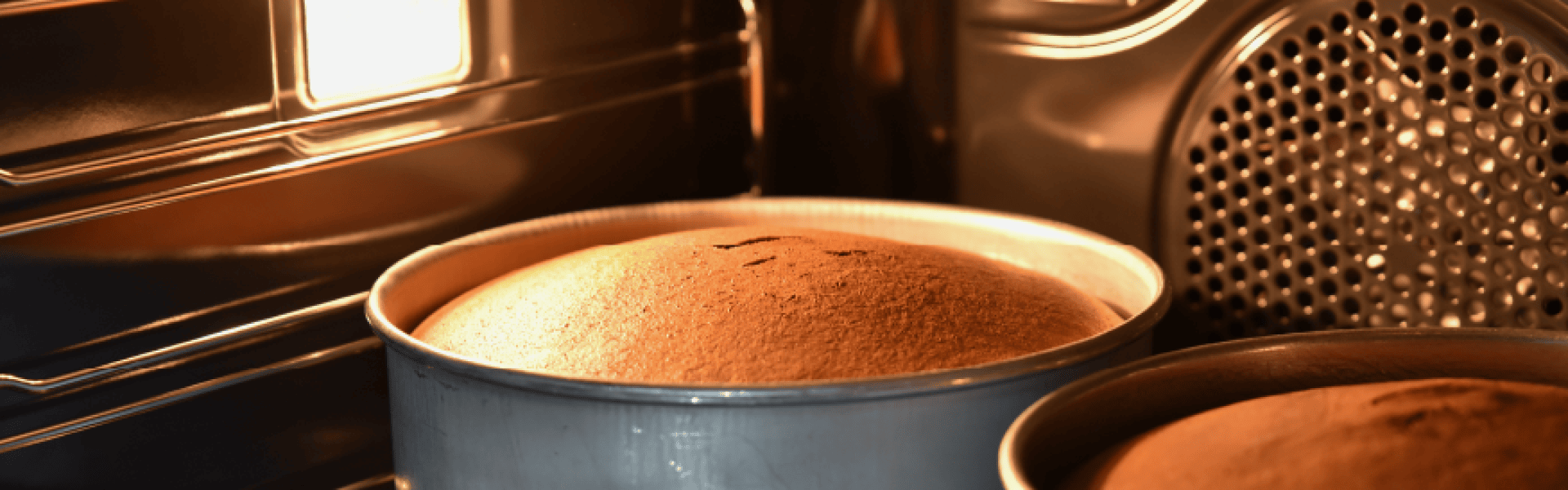 Zwei runde Kuchenformen mit Kuchen, die im Ofen aufgehen, beleuchtet von der Ofenlampe. Die Kuchen sind goldbraun und fast fertig gebacken, wobei ein Kuchen oben leicht gerissen ist.