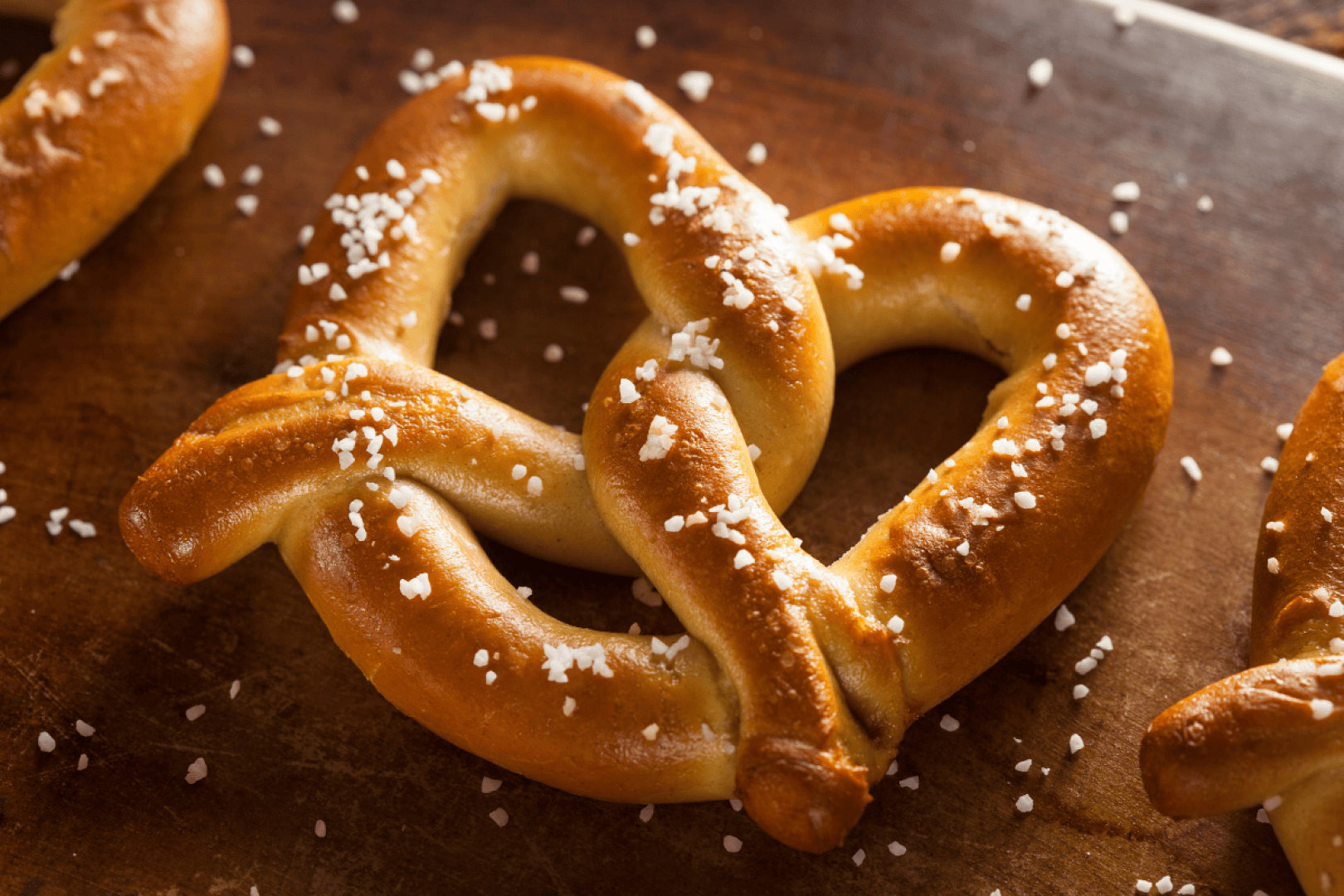 A close-up of a freshly baked soft pretzel sprinkled with coarse salt, resting on a wooden surface.