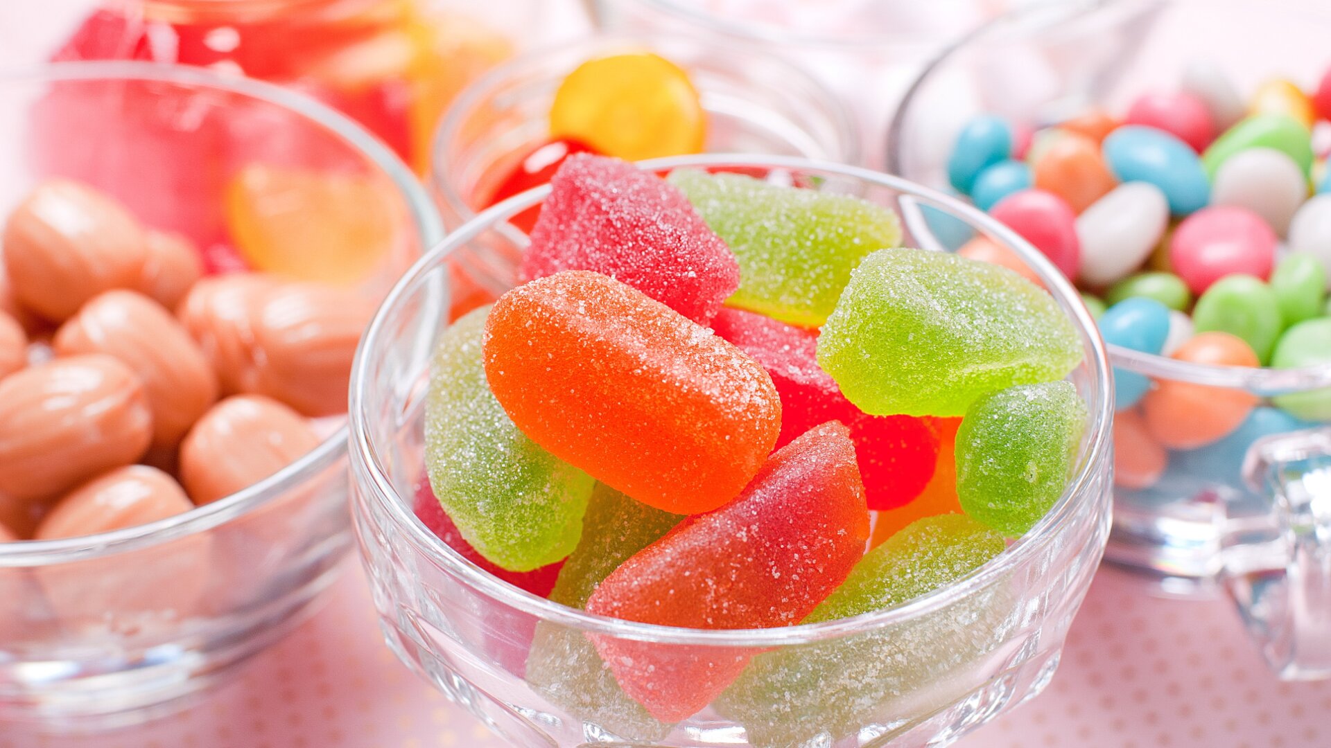Bunt gemischte Bonbons in Glasschalen. Das Bild zeigt mit Zucker überzogene Gummibonbons, Hartkaramellen und Pralinen in verschiedenen leuchtenden Farben, die dicht nebeneinander auf einer hellen Fläche angeordnet sind.