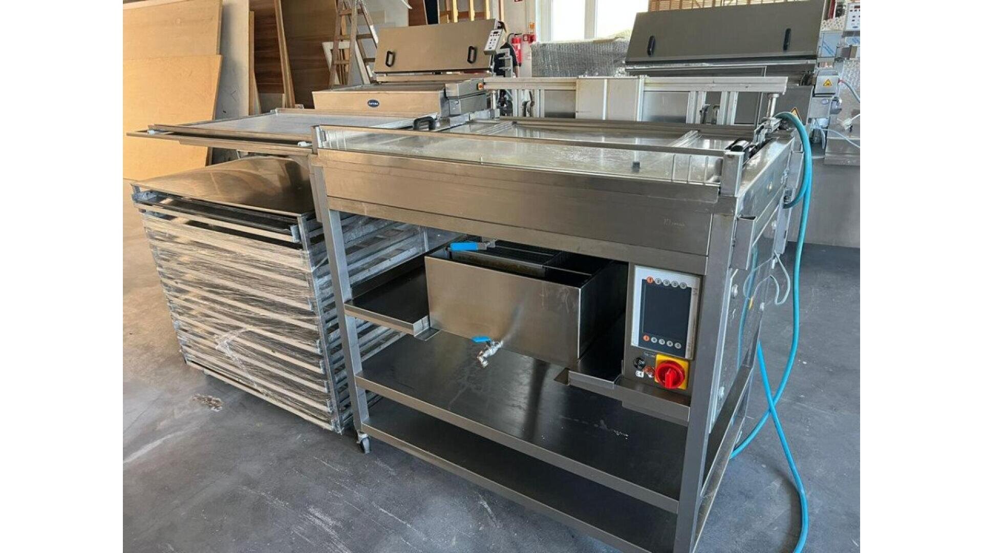 A stainless steel industrial machine with control panel, hoses, sinks, and flat surfaces, situated in a workshop environment with racks and equipment in the background.