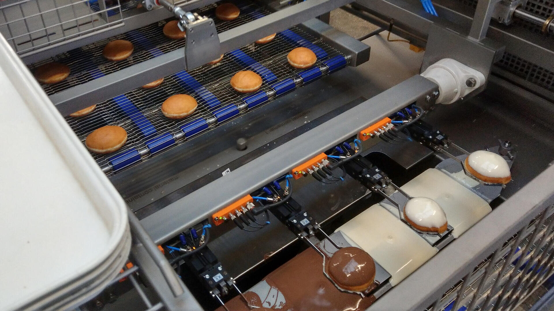 Eine Maschine in einer Fabrik taucht rundes Gebäck in Schokolade und weißen Zuckerguss und legt es dann zum Abkühlen auf ein Förderband, während auf dem oberen Band weiteres, nicht getränktes Gebäck wartet.