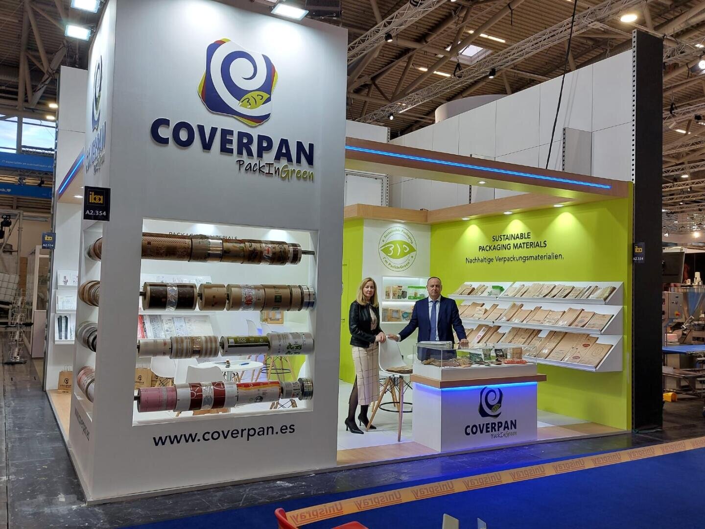 Zwei Personen stehen am COVERPAN-Messestand und präsentieren nachhaltige Verpackungsmaterialien. Muster werden in Regalen und auf einer Theke präsentiert, Markenzeichen und Informationen sind an den Standwänden angebracht.