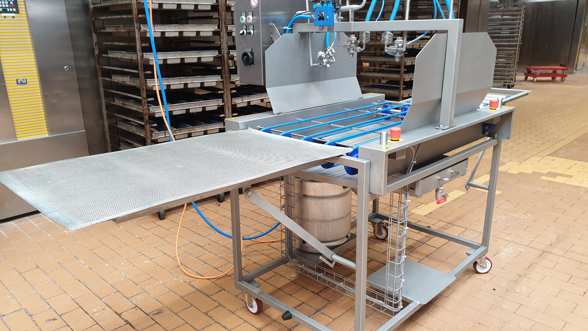 Eine gewerbliche Bäckereimaschine mit Metallgestellen, Förderbändern und Bedienfeldern steht auf Rädern in einer gefliesten Großküche. Unter dem Gerät ist eine große Rührschüssel untergebracht.
