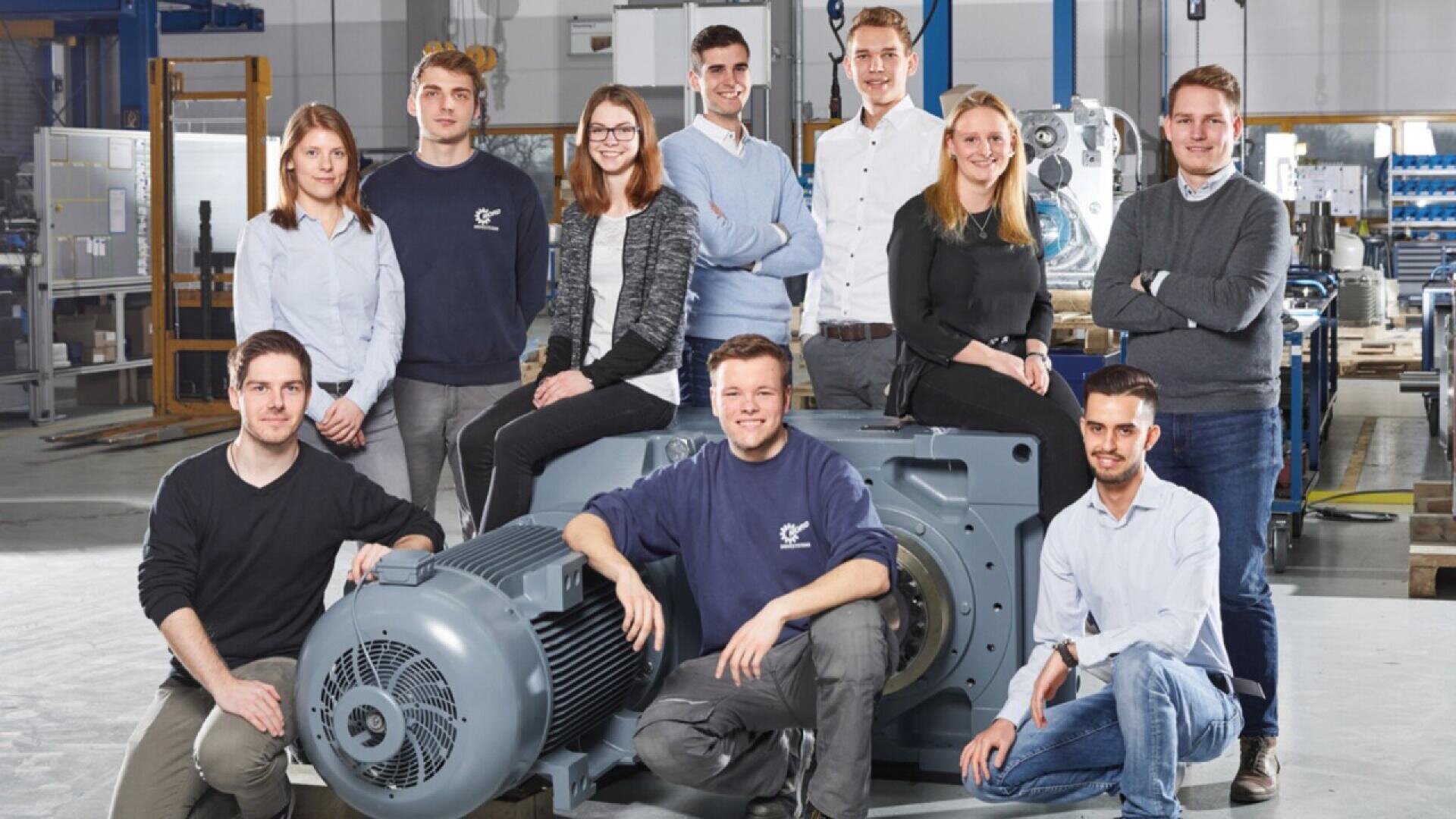 Ten young adults, casually dressed, pose together in an industrial workshop, some sitting and some standing around a large piece of machinery, smiling at the camera.