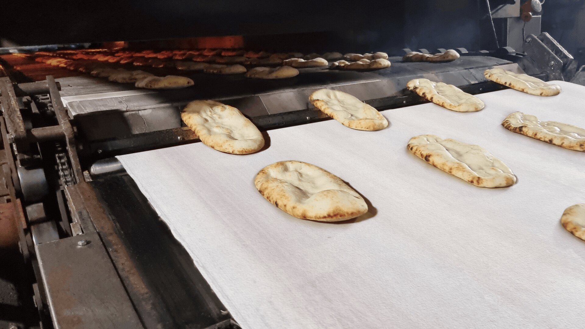 Flatbreads move along a conveyor belt in a commercial bakery oven, with rows of bread baking evenly as they progress through the industrial baking process.