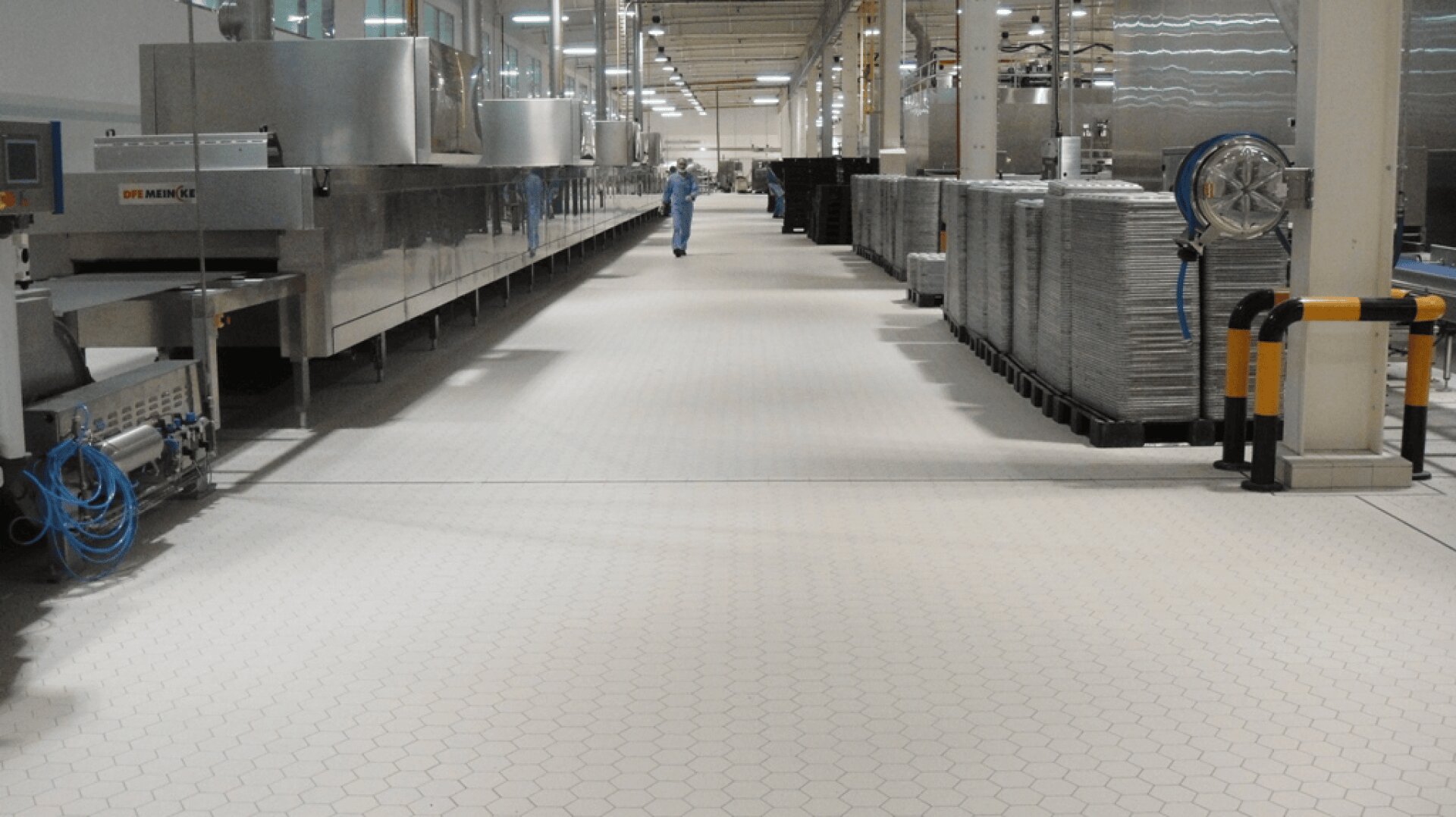 A clean, modern factory interior with hexagonal tile flooring, large industrial machines on the left, stacked metal trays on the right, and a person in protective clothing walking in the distance.