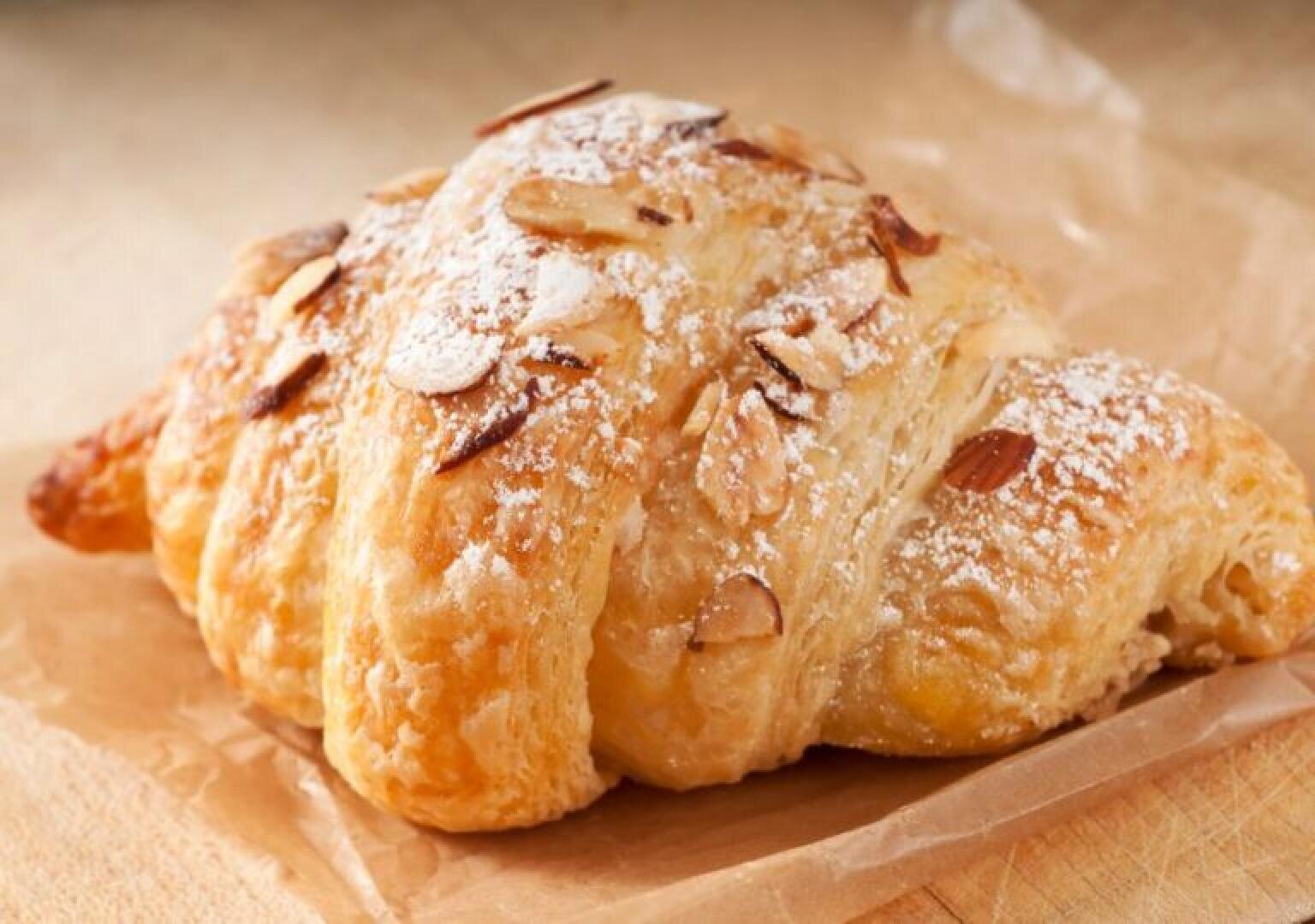 A flaky almond croissant topped with sliced almonds and a dusting of powdered sugar sits on a piece of parchment paper atop a wooden surface.