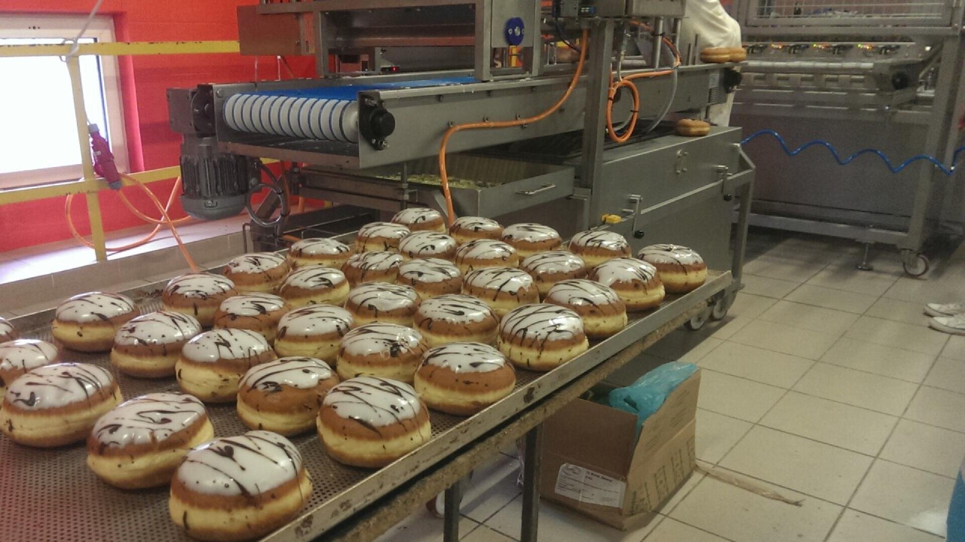 Rows of freshly glazed donuts with chocolate drizzle are arranged on a tray in a bakery production area, with industrial machines and conveyor belts in the background.