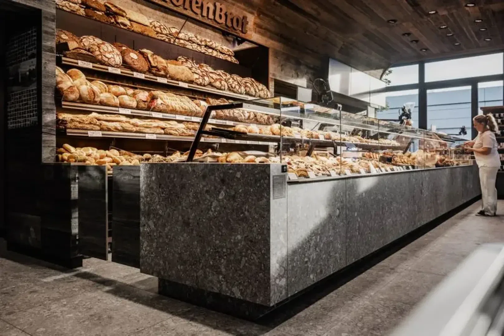 Moderne Bäckerei mit eleganten grauen Theken und Glasvitrinen voller Brot und Gebäck. In den Regalen hinter der Theke sind weitere Brote ausgestellt. Eine Person steht an der Theke, Sonnenlicht fällt hinein.