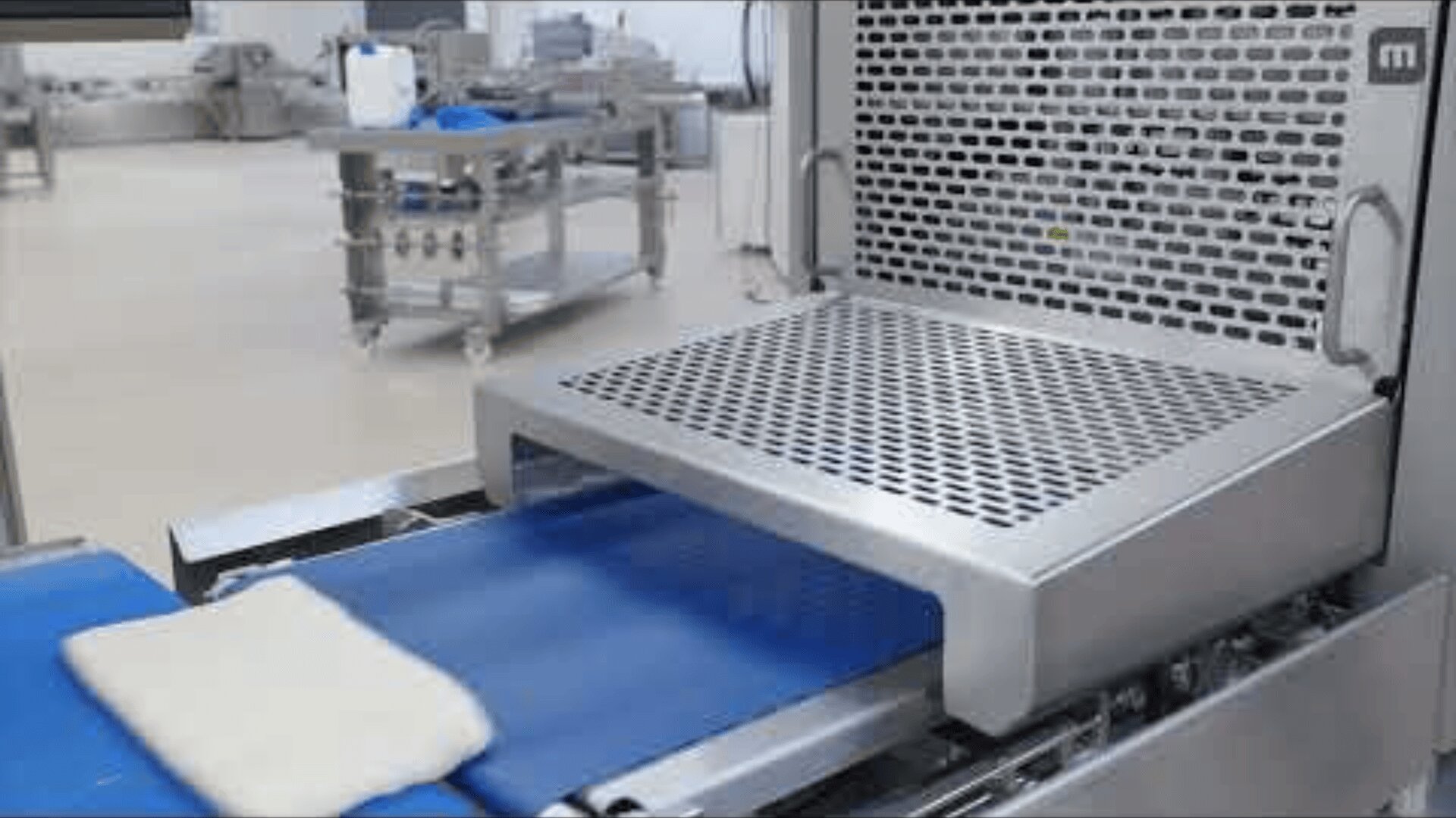 Ein Stück Teig liegt auf einem blauen Förderband und bewegt sich in Richtung einer großen industriellen Lebensmittelverarbeitungsmaschine aus Edelstahl in einer sauberen Fabrikumgebung.