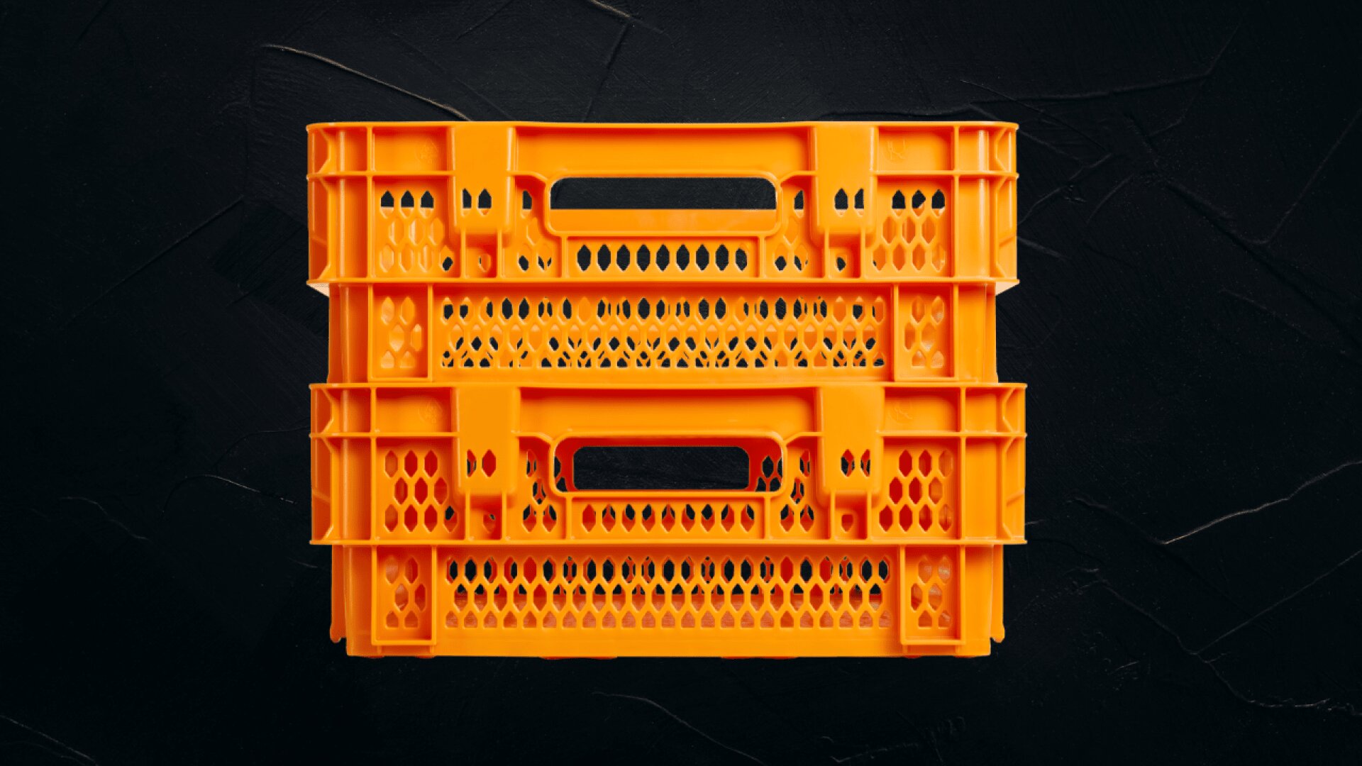Eine leuchtend orangefarbene Kunststoffkiste mit perforierten Seiten und Griffen ist auf einer dunkel strukturierten Oberfläche gestapelt.