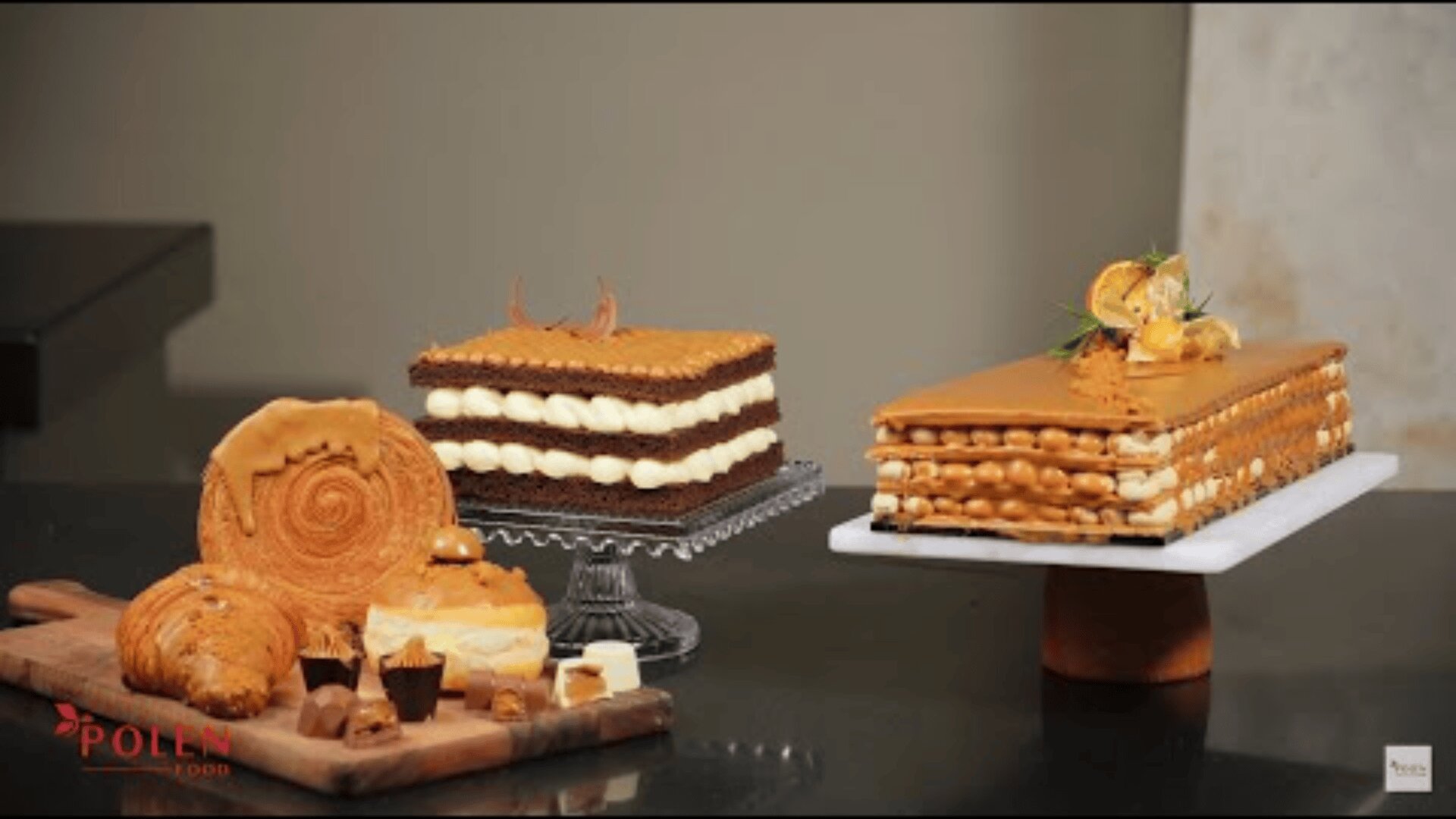 Three elegant cakes with layers of cream are displayed on stands, accompanied by assorted pastries, including a spiral roll and muffins, all arranged on a black tabletop against a neutral background.