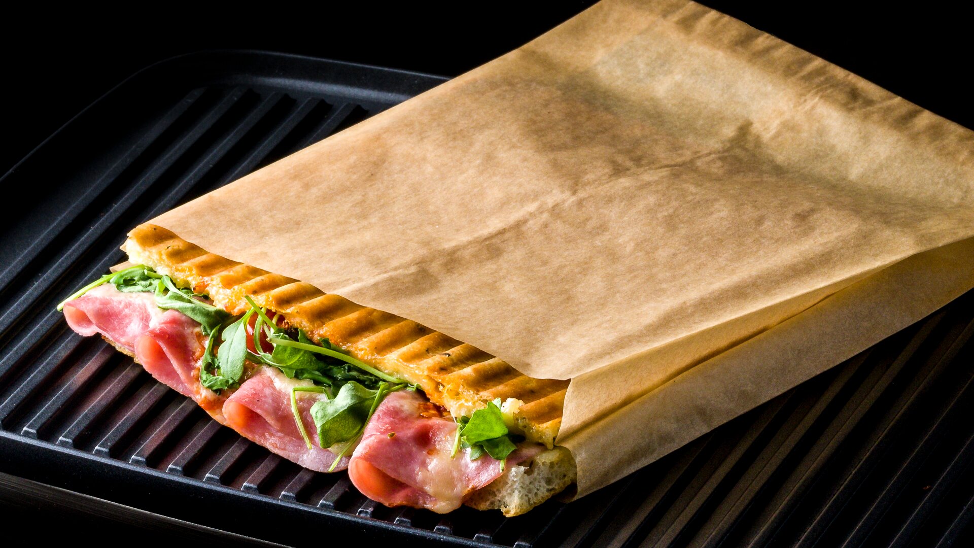 A grilled panini sandwich filled with ham and fresh greens is partially wrapped in brown paper and placed on a black grill pan.