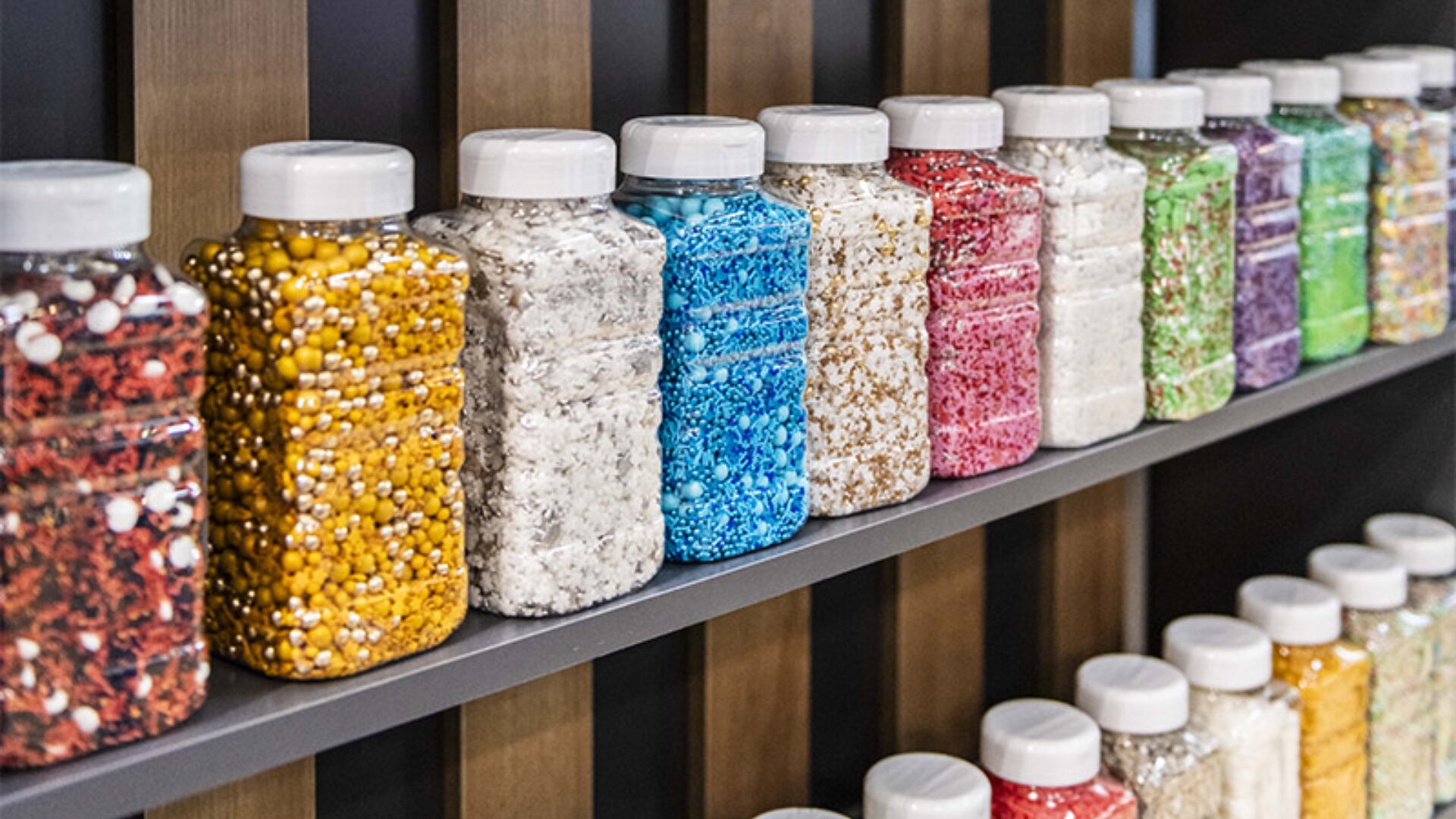 Klare Plastikdosen mit Streuseln in verschiedenen Farben und Formen sind auf dunkelgrauen Regalen vor einem hölzernen Hintergrund fein säuberlich aufgereiht.