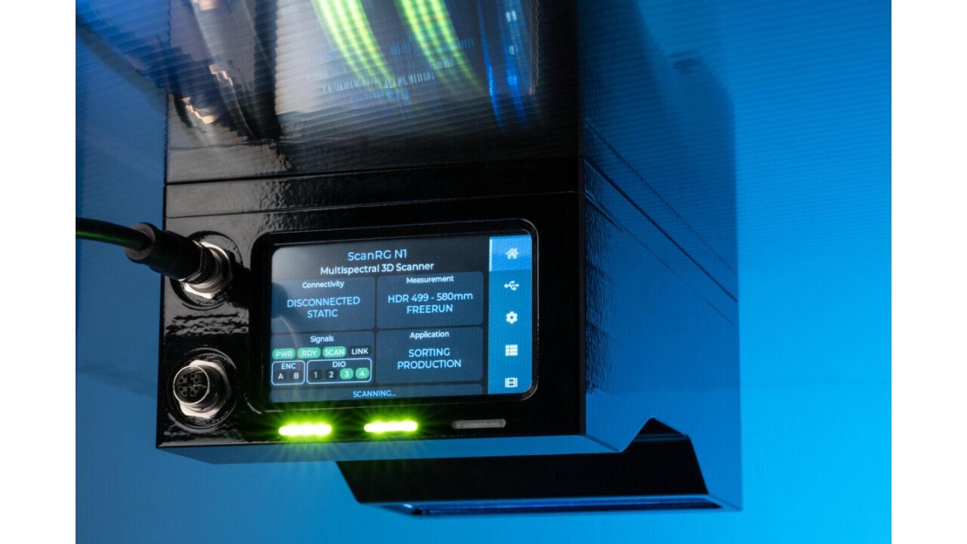 A close-up of a black multispectral 3D scanner device with a display screen showing connectivity and signal status, illuminated by blue light. Green indicator lights and connected cables are visible on the left side.