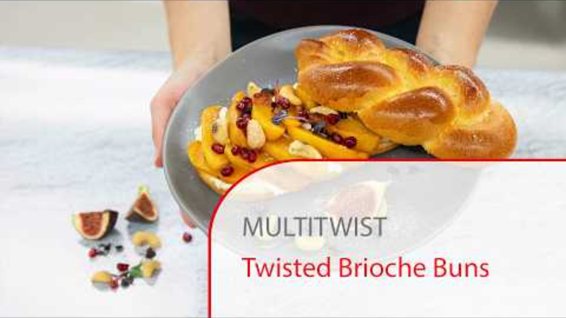 A person holds a plate with a twisted brioche bun, garnished with fruit and nuts. Sliced figs and more fruit are scattered on the table. Text on the image reads “MULTITWIST Twisted Brioche Buns.”.