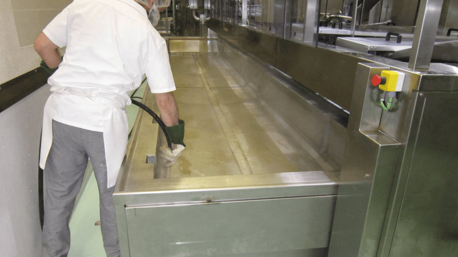 A person dressed in a white uniform and gloves is cleaning or washing a large industrial metal sink or basin with a hose in a commercial kitchen or food processing area.