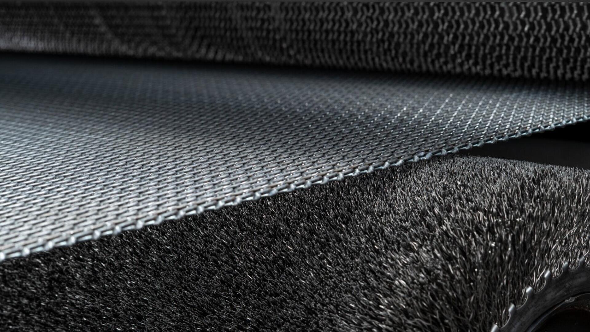 Close-up of an industrial conveyor belt with a textured, mesh-like surface and a soft, fibrous material beneath, showing detailed patterns and material contrast.