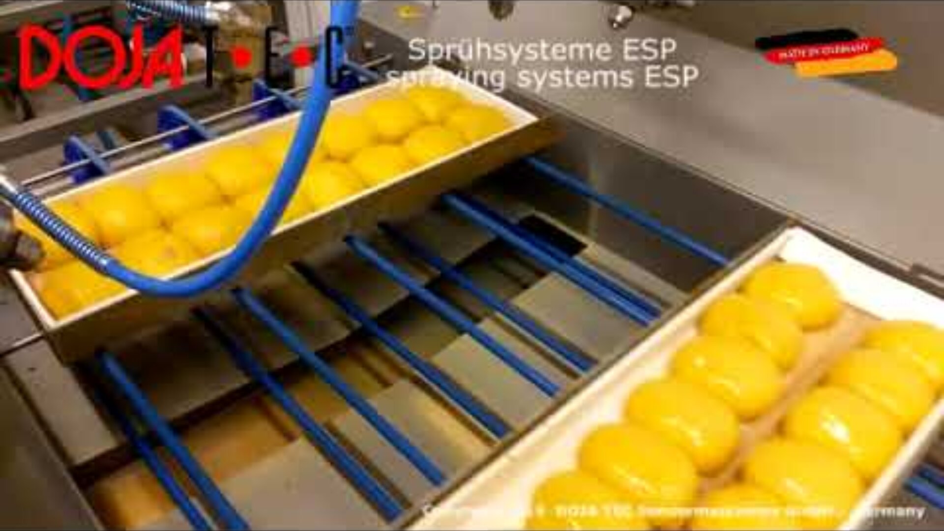 Gelbe Backwaren sind in Reihen auf Tabletts angeordnet, die in einer Fabrik über ein Förderband laufen. Blaue Rohre und Maschinen tragen eine Spritzschicht auf. Der Text in Deutsch und Englisch lautet: Sprühsysteme ESP, spraying systems ESP.