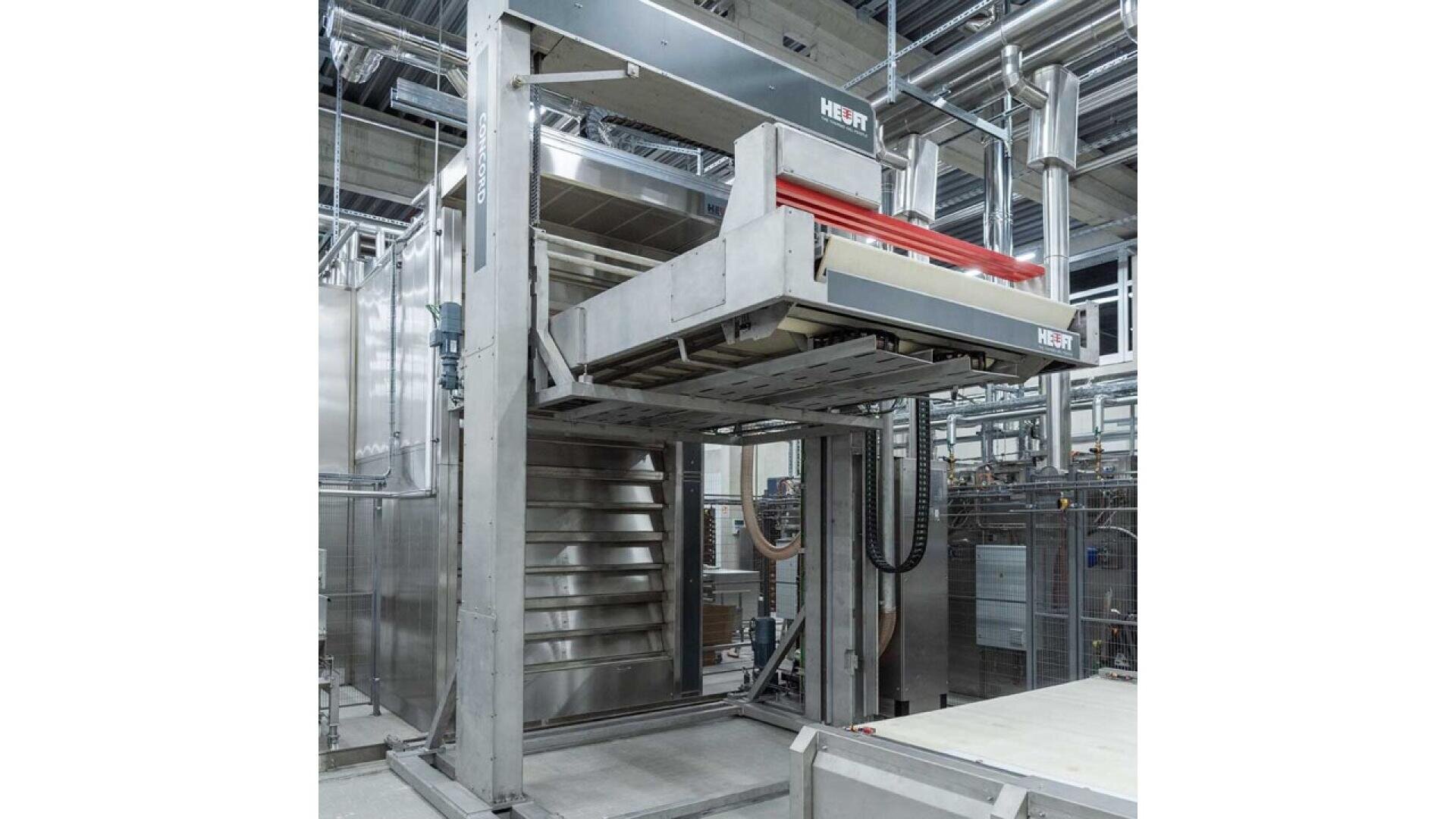 A large, industrial bakery oven with multiple baking chambers and a mechanical loading system is shown in a factory setting with exposed pipes and cables overhead.