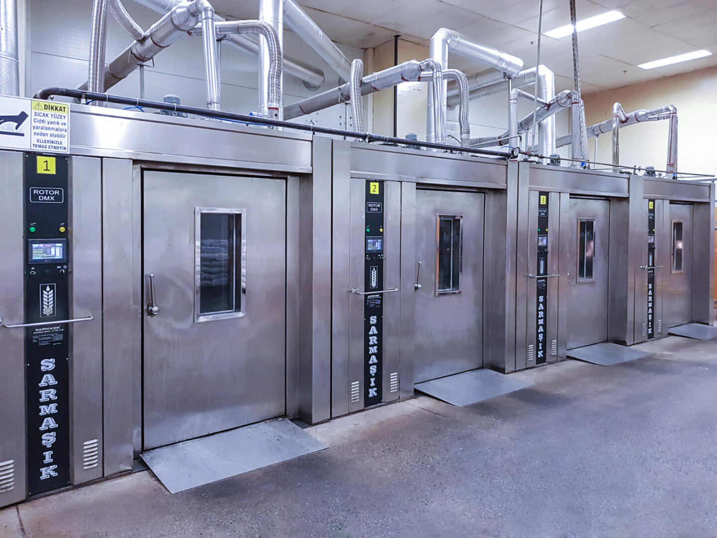 Four large industrial stainless steel ovens with digital control panels stand side by side in a clean, well-lit facility. Pipes and vents run overhead, and the floor is smooth and uncluttered.