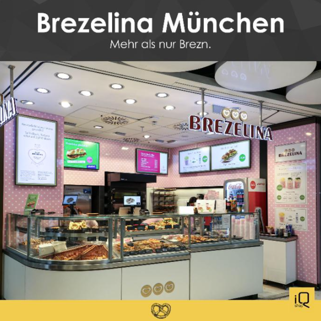 Ein Brezelina-Laden in München mit einer Glasvitrine mit Backwaren, rosa-weißem Dekor, Menütafeln und einem Schild mit der Aufschrift "Brezelina München. Mehr als nur Brezn.".