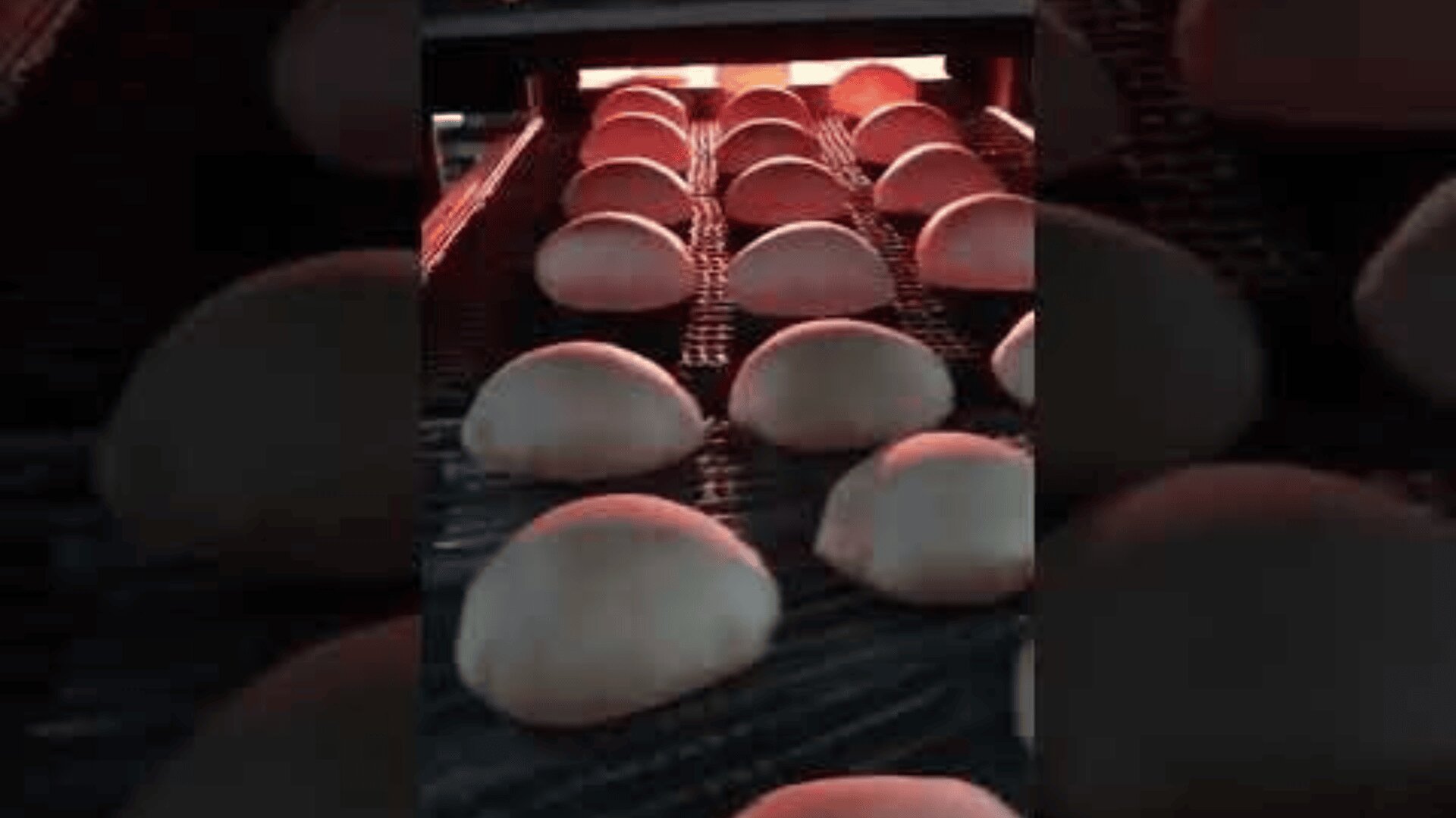 In einer Bäckerei oder Fabrik laufen Reihen von runden Brötchen auf einem Förderband unter einer Wärmequelle.