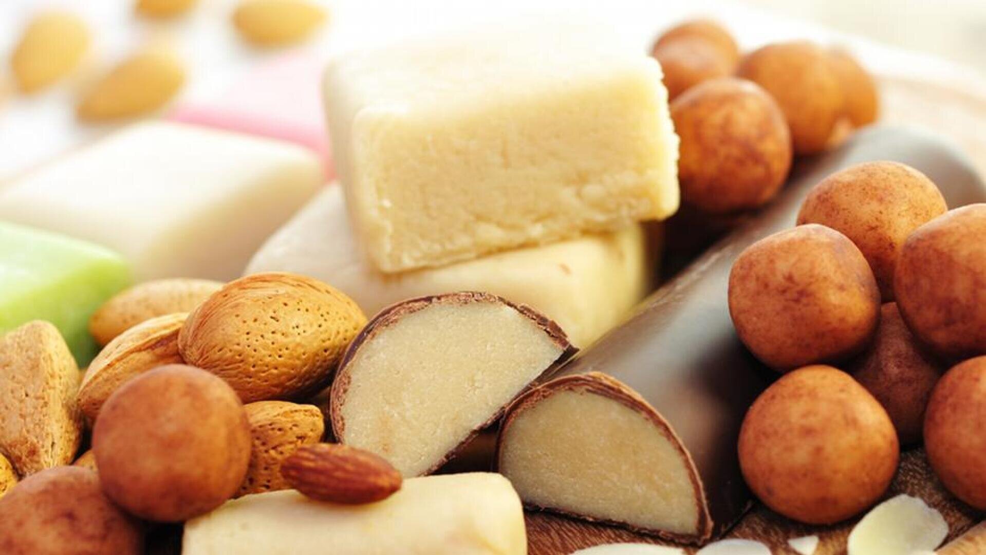 Eine Auswahl an Marzipanbonbons, darunter mandelförmige Stücke, schokoliertes Marzipan, ganze Mandeln und verschiedene Marzipanblöcke in unterschiedlichen Farben, dicht an dicht angeordnet.