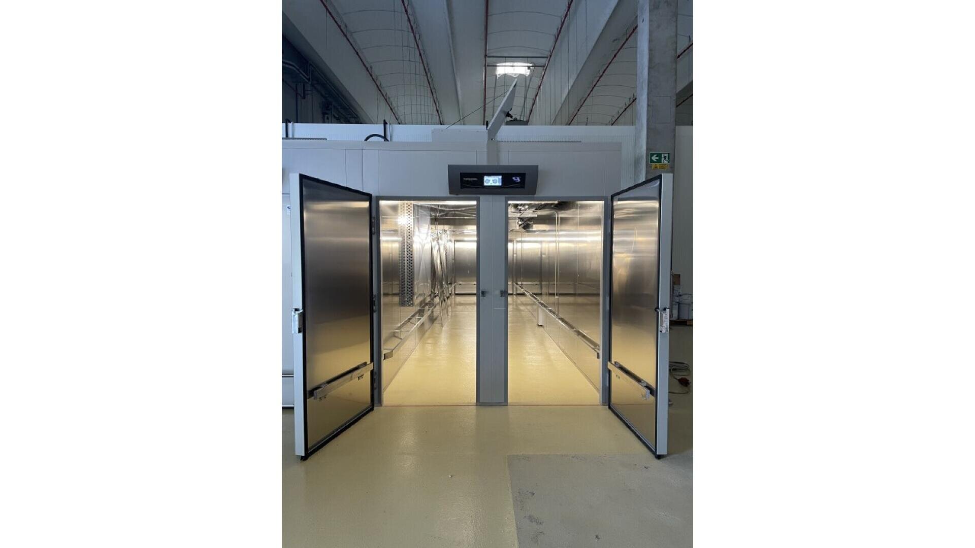 Ein hell erleuchteter industrieller Kühlraum mit offenen doppelten Metalltüren, die den Blick auf Edelstahlregale im Inneren freigeben. Der Raum hat ein digitales Bedienfeld über dem Eingang und einen sauberen, glatten Boden.