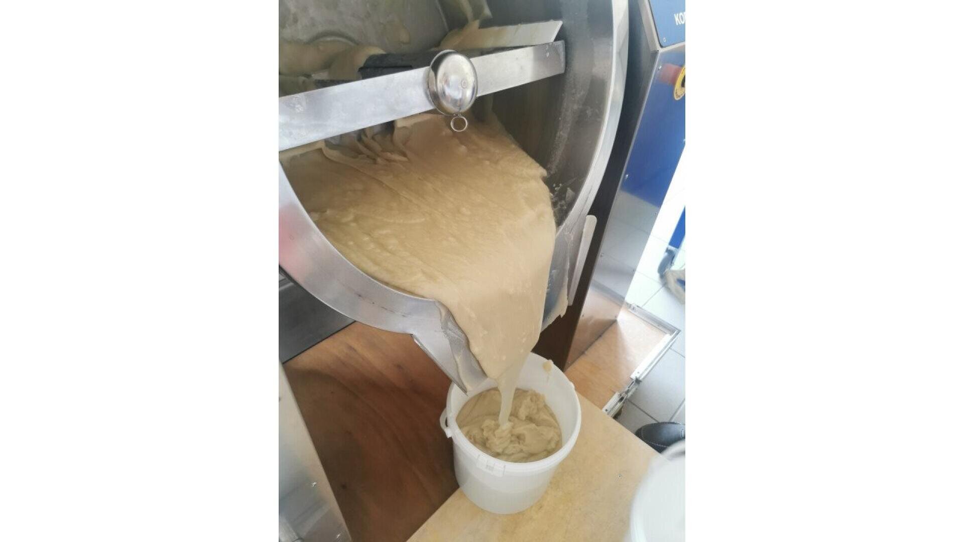 Thick, creamy batter pours from a large industrial mixing machine into a white plastic bucket below in a commercial kitchen setting.