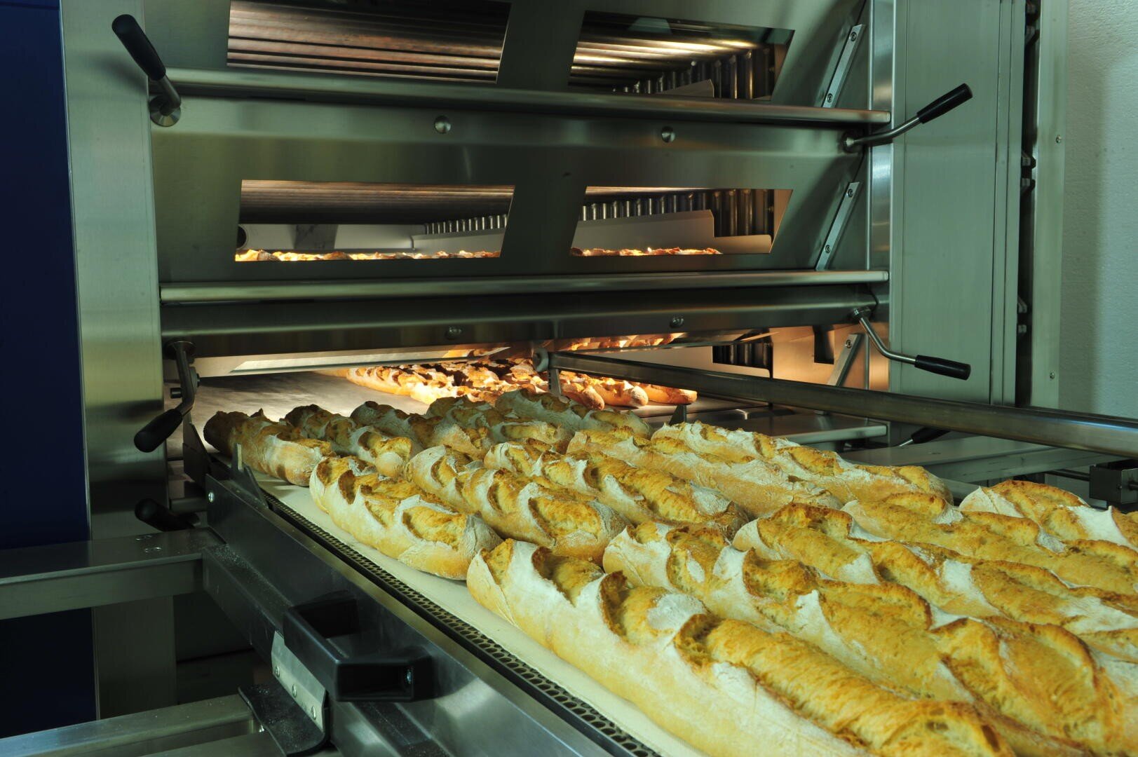 Frisch gebackene Baguettes kommen in einer Großbäckerei auf einem Fließband aus einem Industrieofen.