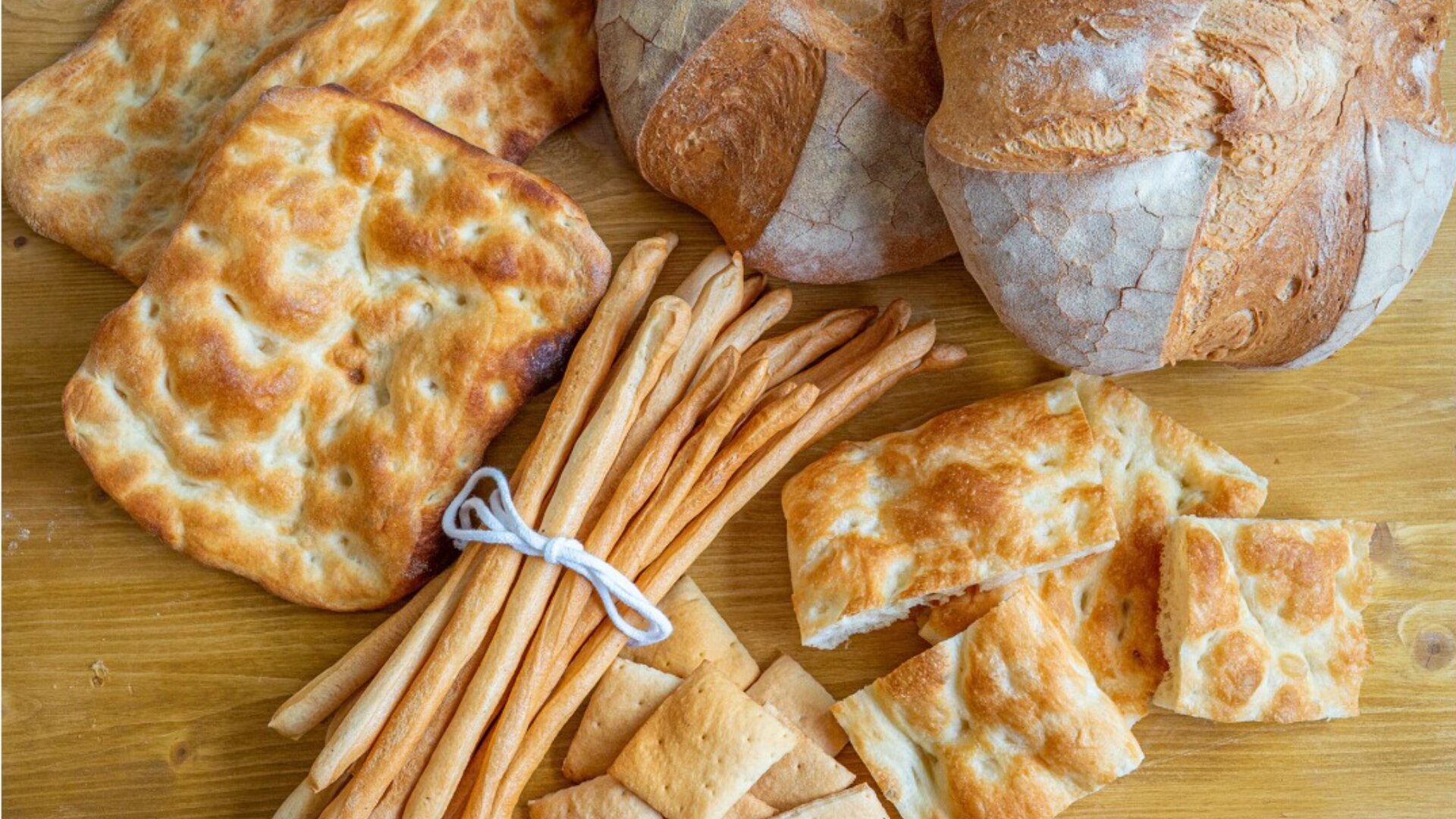 Eine Auswahl italienischer Brotsorten auf einer hölzernen Unterlage, darunter runde Brote, quadratische Focaccia-Stücke, dünne, mit Schnur gebundene Brotstangen und kleine rechteckige Cracker.