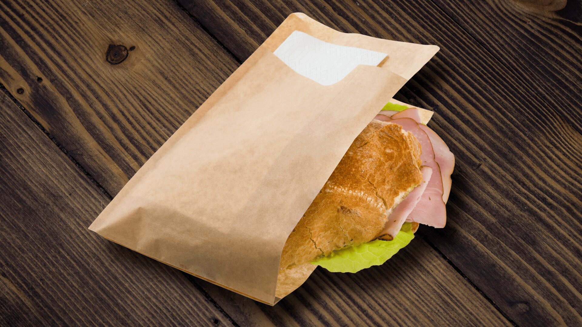 A ham and lettuce sandwich partially wrapped in a brown paper bag sits on a rustic wooden table.