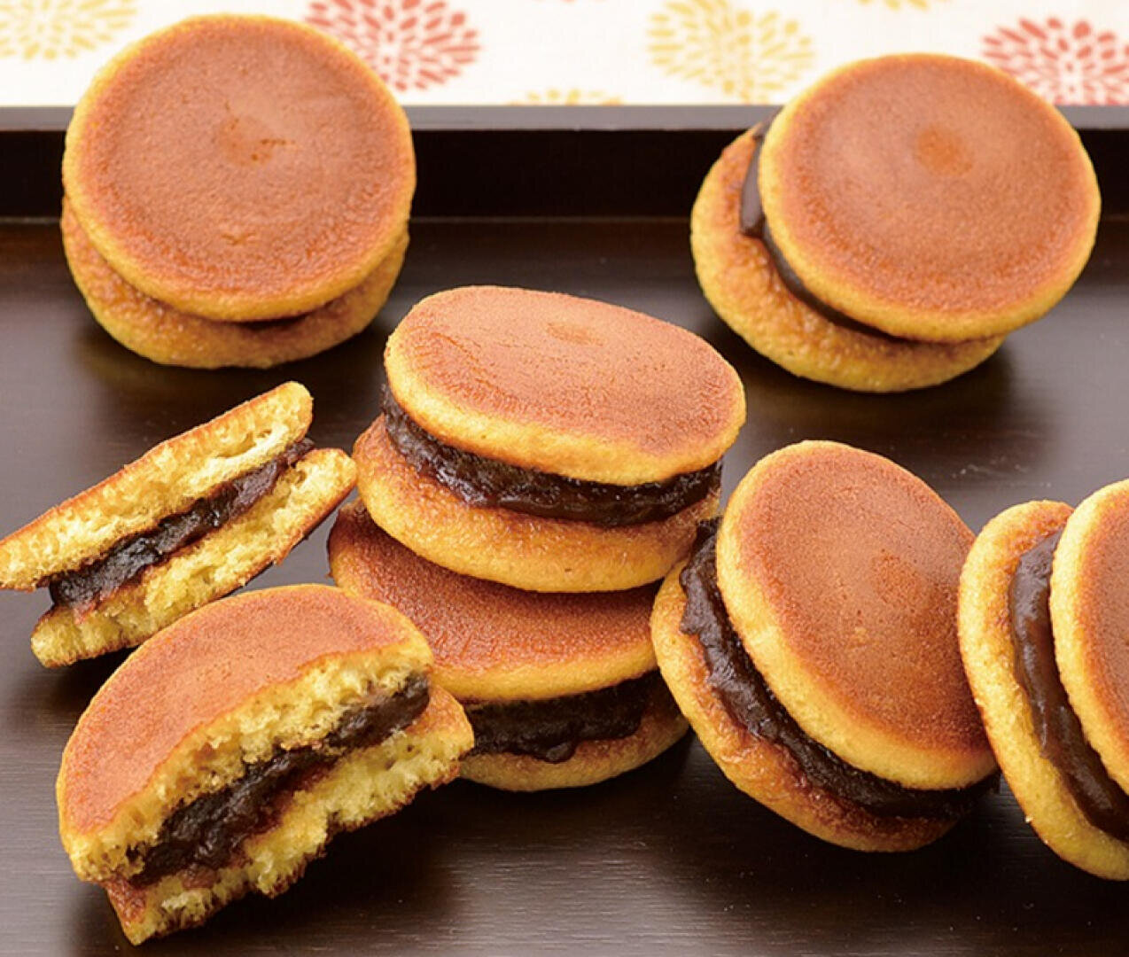 Mehrere Dorayaki, eine japanische Süßigkeit aus zwei runden Pfannkuchen, gefüllt mit süßer roter Bohnenpaste, liegen auf einem dunklen Tablett. Einige sind ganz, andere sind aufgeschnitten, sodass die Füllung sichtbar ist.