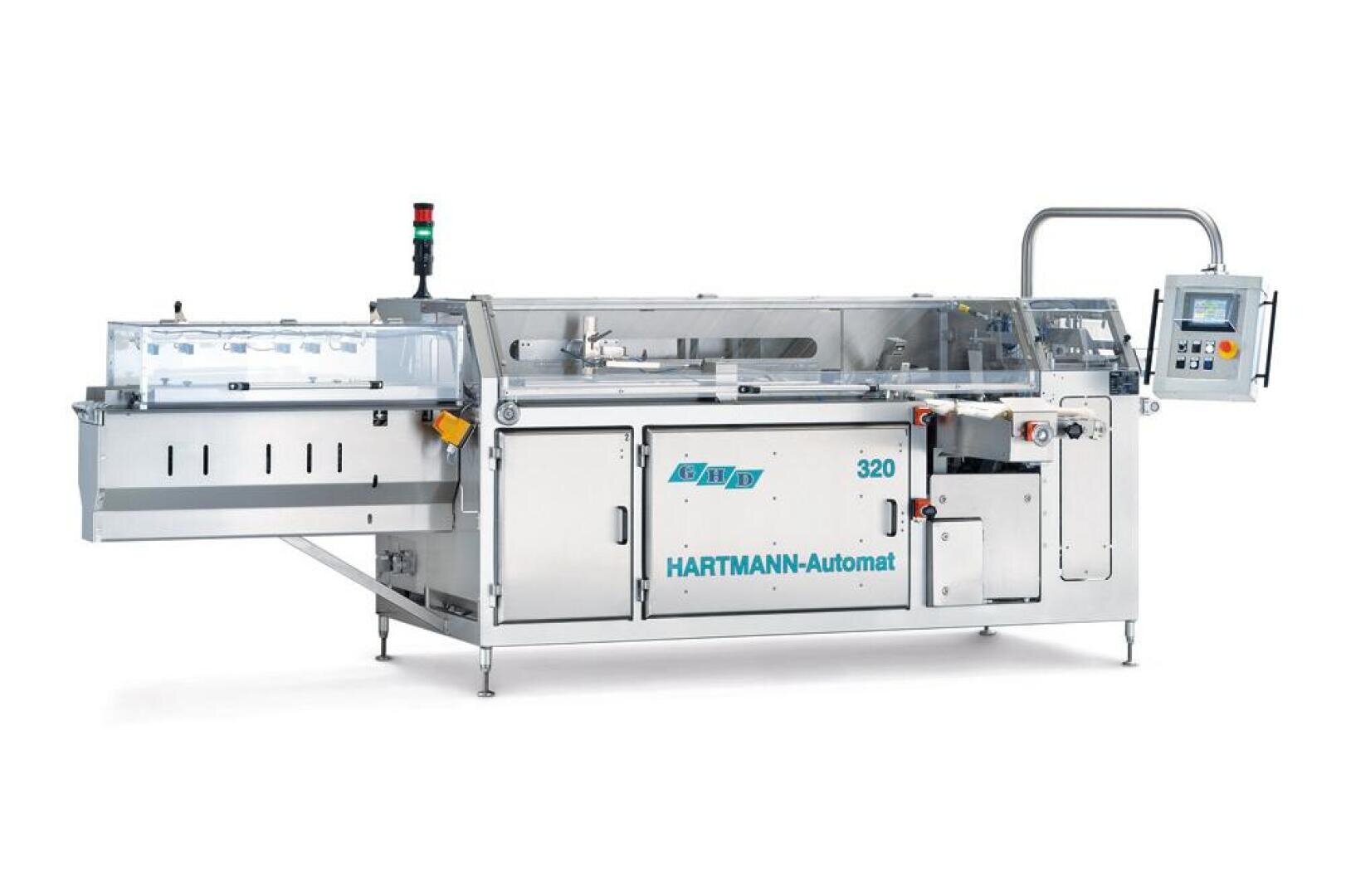 Eine Hartmann-Automat 320 Verpackungsmaschine aus rostfreiem Stahl mit transparenter Abdeckung, Bedienpult, Not-Aus-Taster und verschiedenen Förderstrecken, konzipiert für die automatisierte industrielle Verpackung.