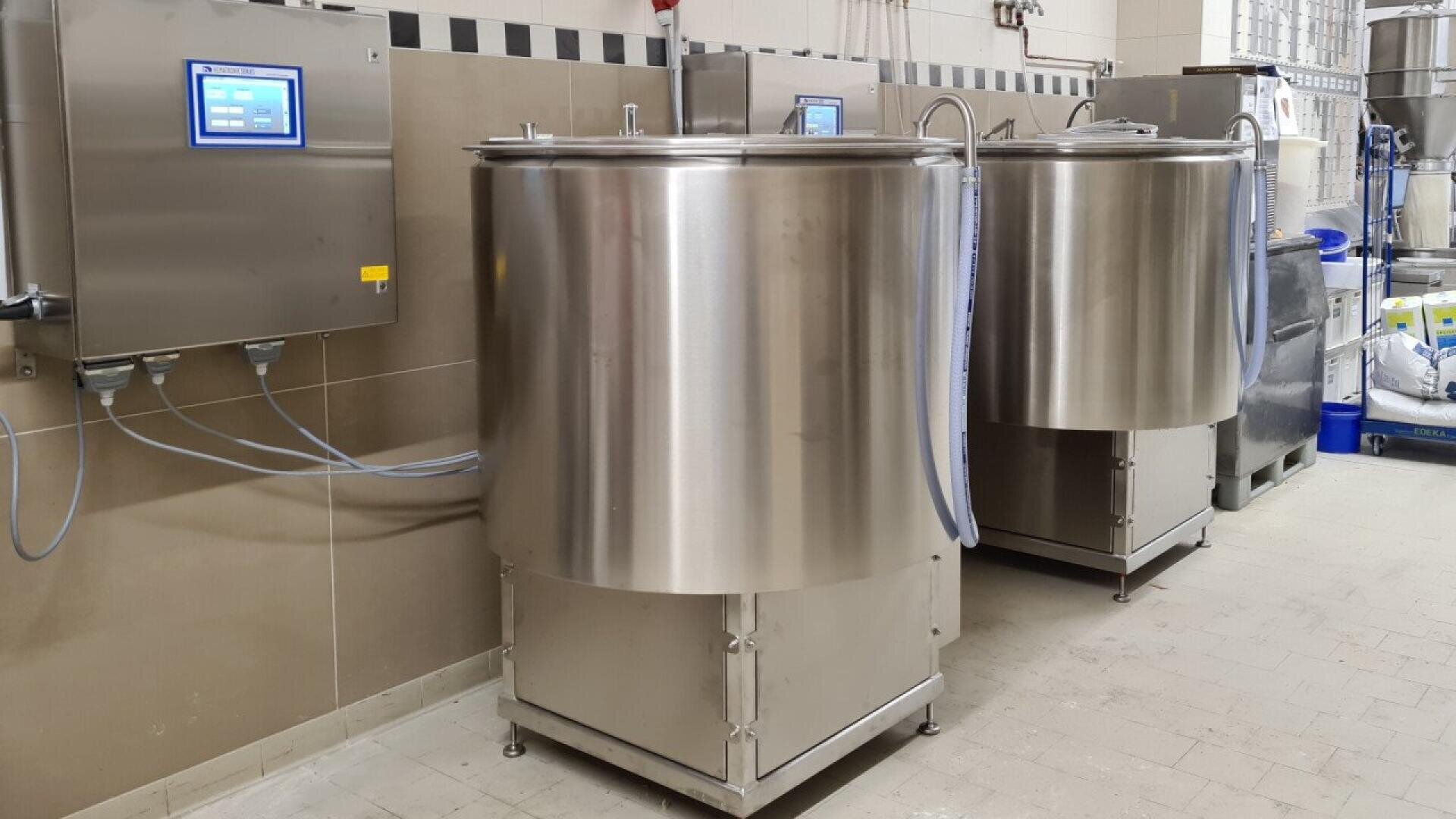 Two large stainless steel industrial tanks with digital control panels are positioned against a tiled wall in a clean facility, possibly a dairy or food processing plant. Blue hoses connect the tanks to the panels.