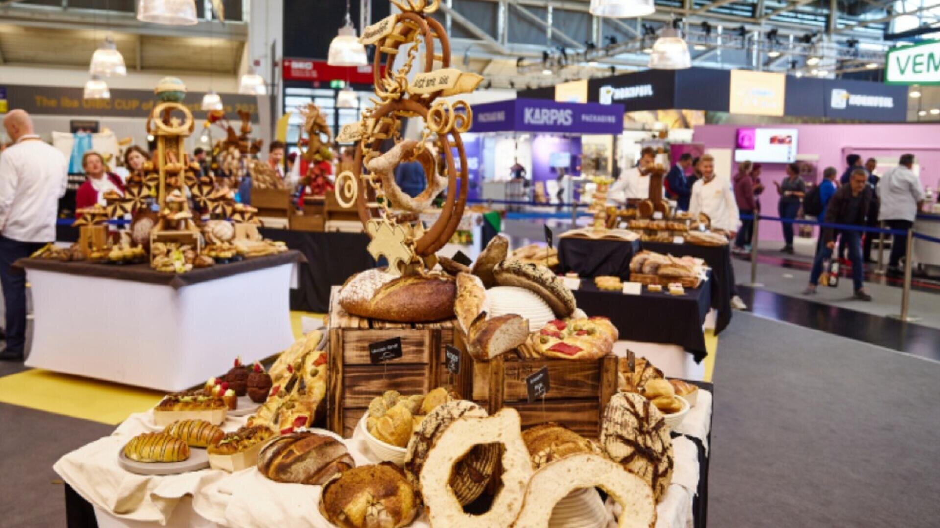 A display of assorted artisan breads and pastries arranged on wooden crates and cloth, with intricate bread sculptures at a bustling bakery or culinary competition event, people and booths visible in the background.