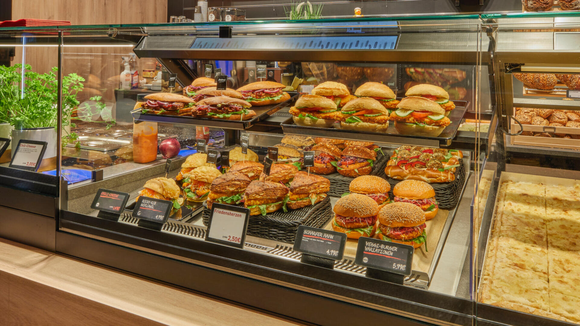 Eine Vitrine in einem Feinkostladen oder Café, die mit verschiedenen Sandwiches, Burgern und Paninis gefüllt ist, mit frischen Zutaten und beschrifteten Preisschildern, die überall in der Vitrine sichtbar sind.